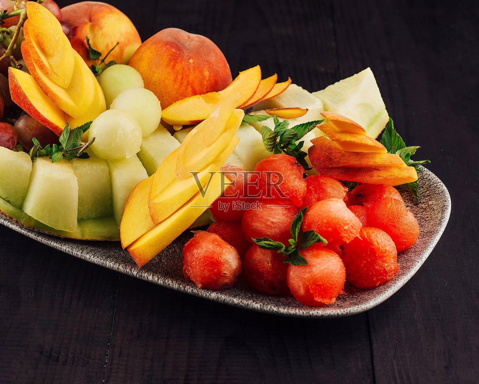 盛满新鲜西瓜、哈密瓜、桃子和葡萄的椭圆盘子放在深色木桌上照片摄影图片