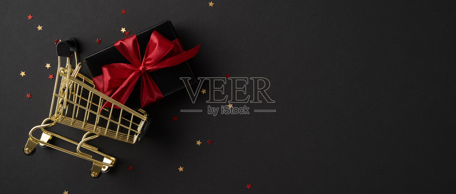 金色购物车上放着一个红丝带礼品盒，周围散落着小星星彩纸，象征着节日购物。照片摄影图片