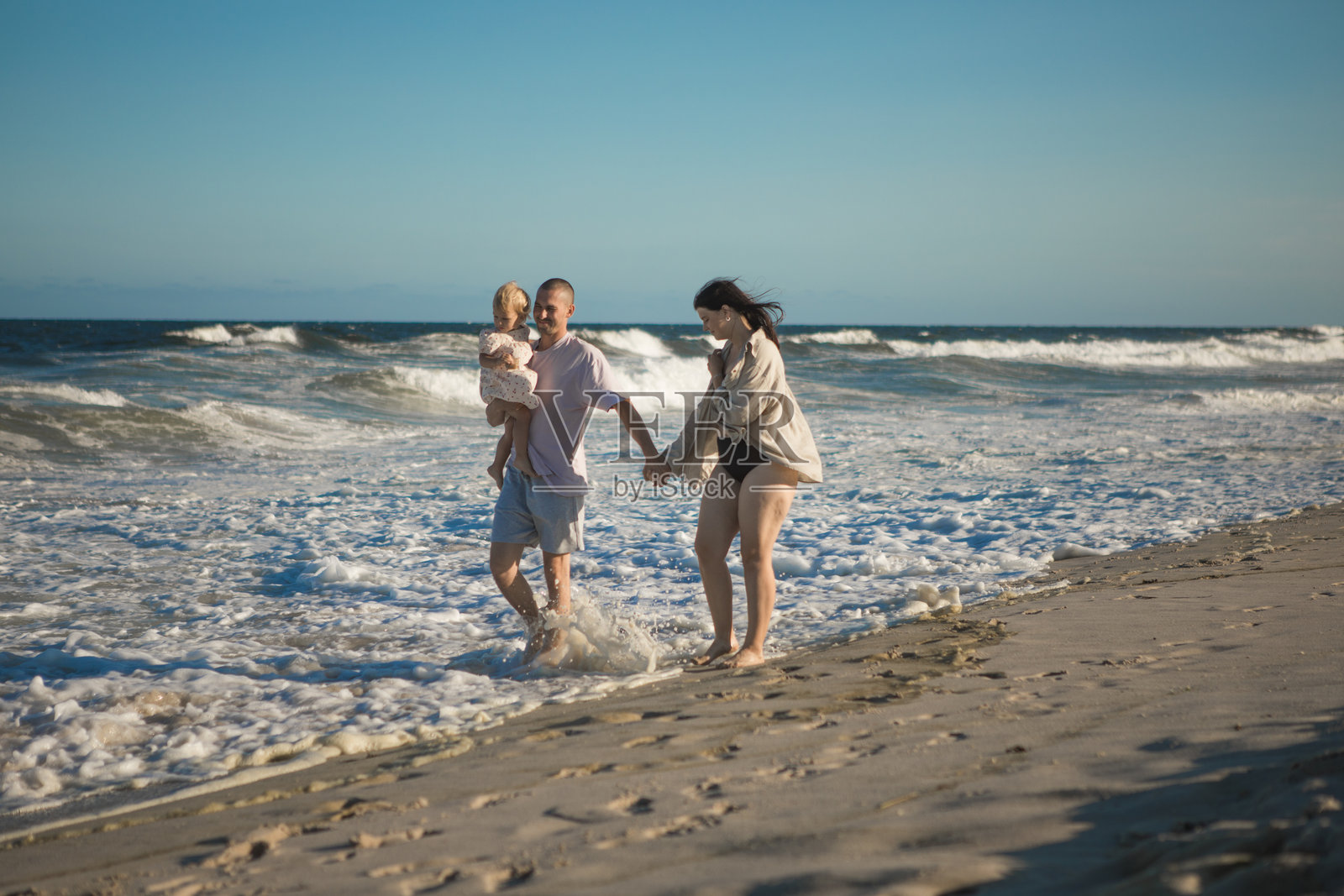 一位年轻的父母牵着她的小女儿的手，一起沿着海洋走向日落。照片摄影图片