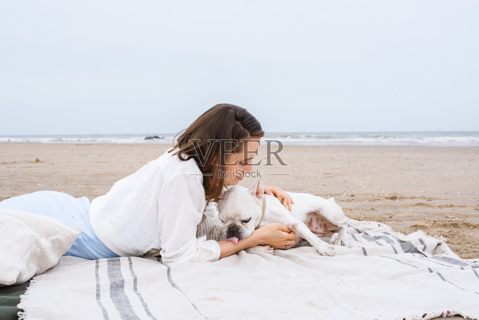 一位美丽的女性和她的法国斗牛犬伴侣在海滩的毯子上共度时光。照片摄影图片