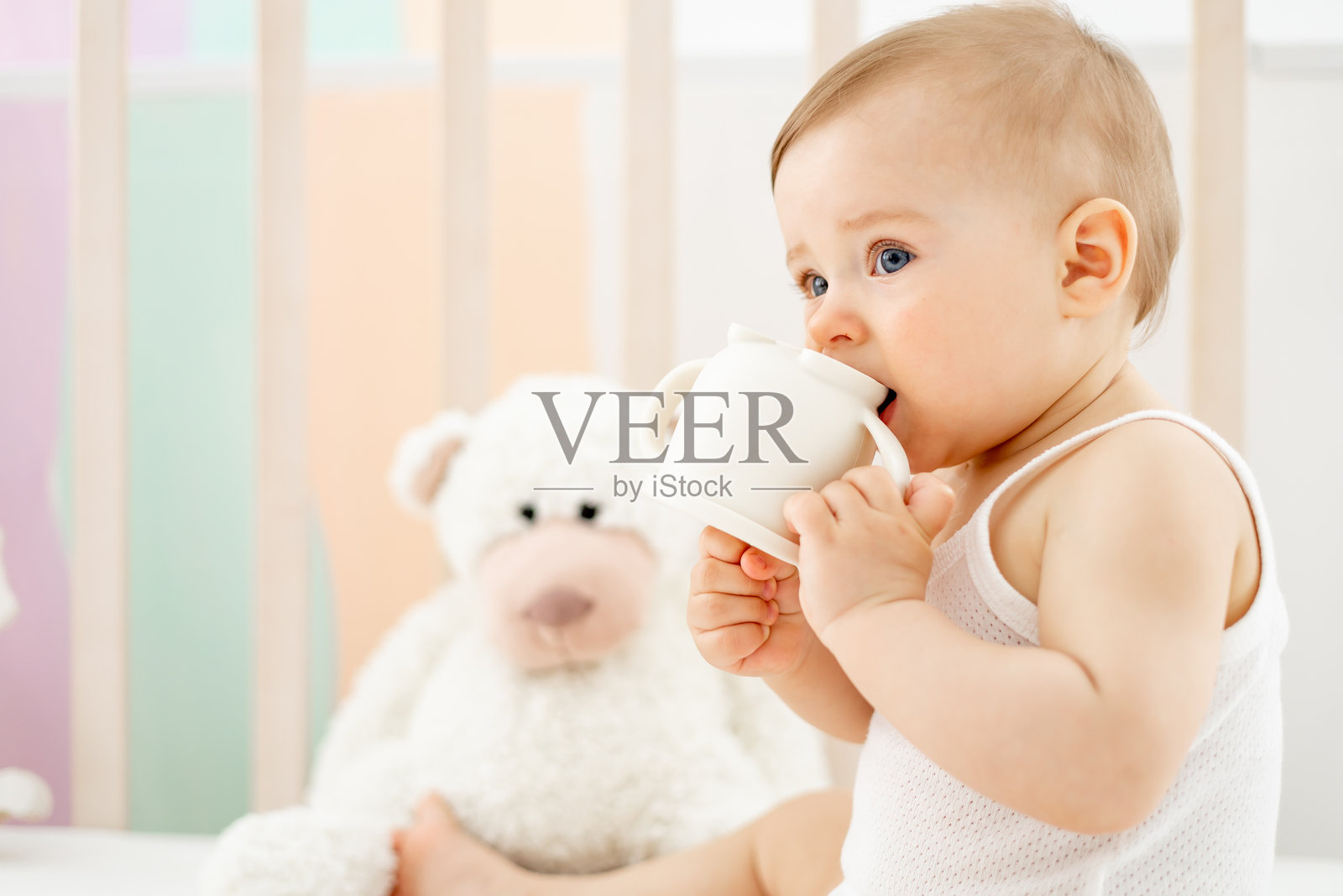 一个小女孩在儿童房的婴儿床上，手里拿着奶瓶或饮水杯，穿着白色连体衣，面带微笑或大笑，十分可爱搞笑，生活方式。照片摄影图片