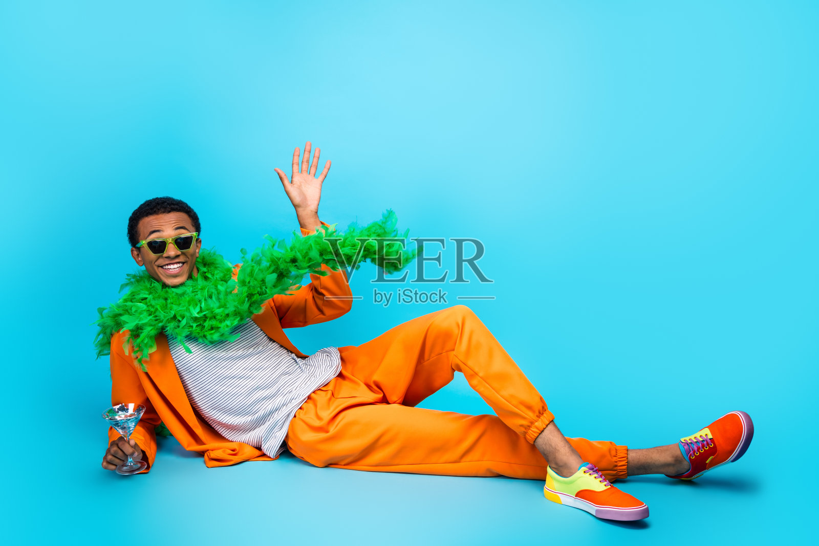 穿着时尚潮流西装的疯狂家伙躺着，手持鸡尾酒杯，挥手致意，背景为蓝色。照片摄影图片