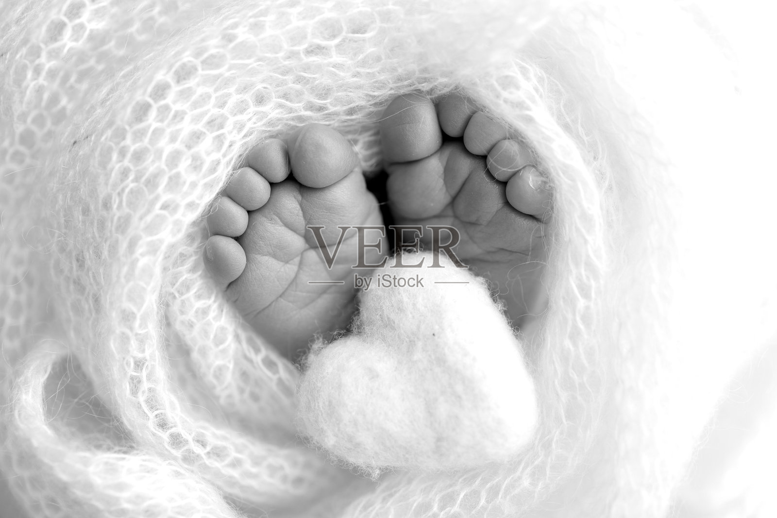 新生儿的小脚丫。包裹在羊毛毯子里的柔软新生儿脚。特写新生儿的脚趾、脚跟和脚。婴儿腿间的编织心形。工作室微距摄影。黑白色调。照片摄影图片