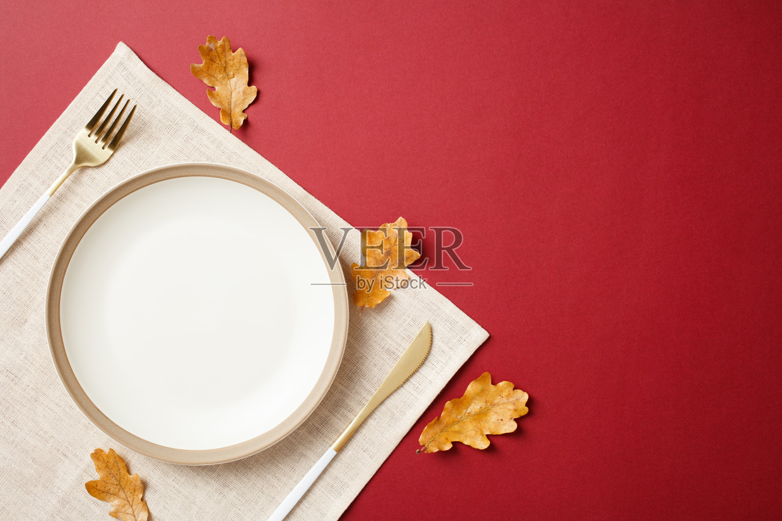 优雅的秋季餐桌布置，白色空盘、金色餐具和散落的秋叶在红色背景上。感恩节用餐概念照片摄影图片