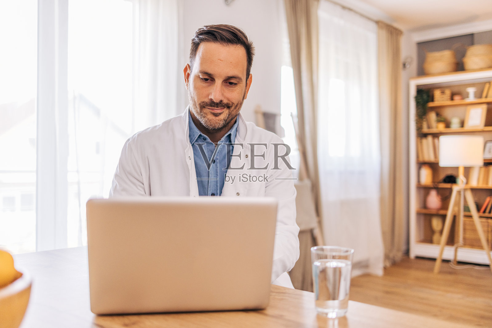 一位中年、开朗的男医生在家中的客厅使用笔记本电脑。照片摄影图片