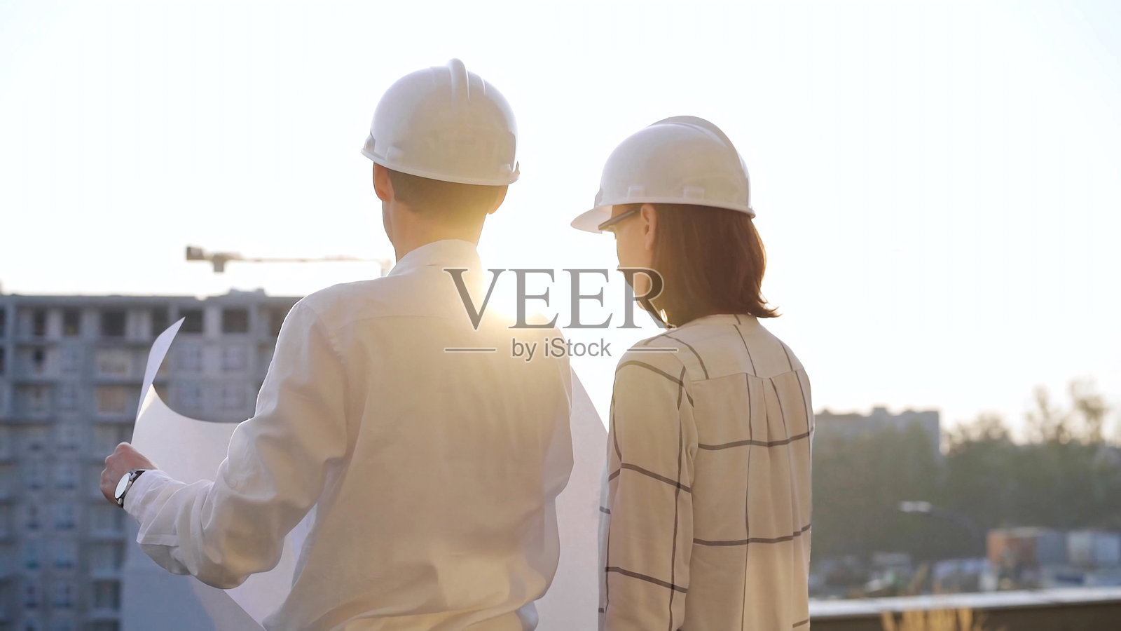 一对男女人建筑师和工程师，戴着安全眼镜和安全帽，在日落时分的建筑工地上查看蓝图，背影。建筑概念照片摄影图片