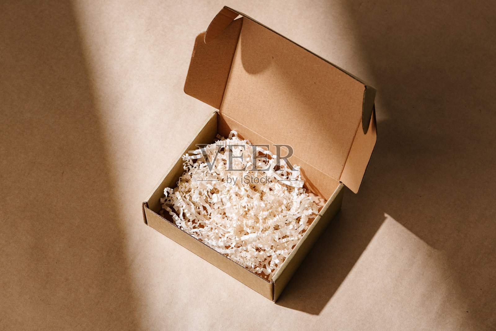 打开的纸箱，里面有碎纸包装，放在柔和自然光照射下的浅棕色表面上。照片摄影图片