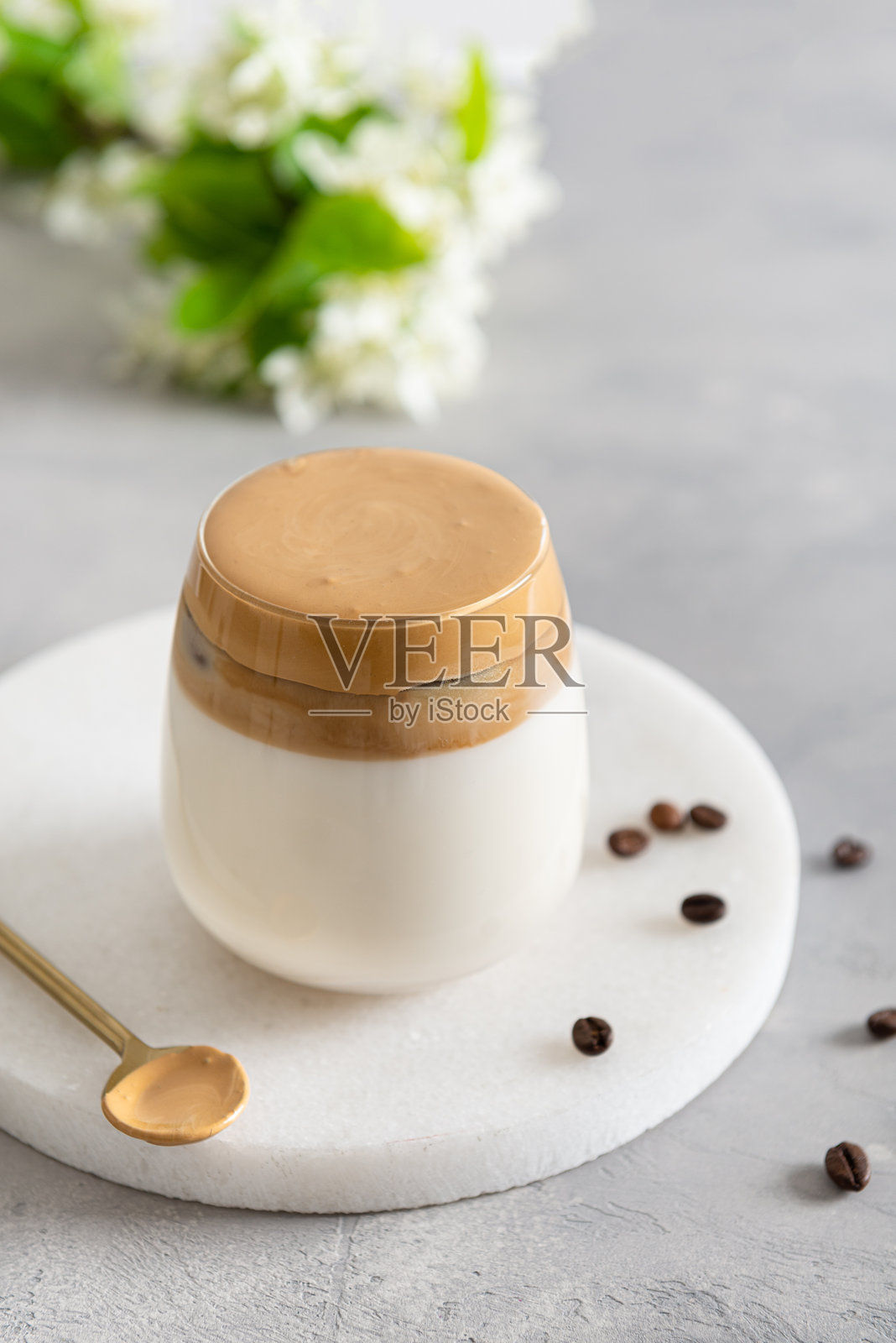 自制的达尔戈纳咖啡，盛在玻璃杯中，旁边有咖啡豆，放在白色桌子上。这款流行的韩国饮品拿铁，顶部是速溶咖啡泡沫。是在隔离和自我隔离期间创造的新饮品。照片摄影图片