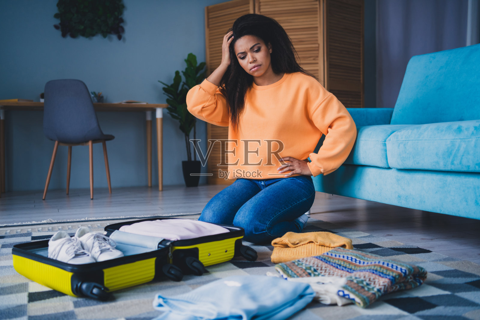 全身照：一位迷人的年轻女性坐在地板上，打包行李箱，神情困惑，穿着橙色服装，享受温馨的日光家庭室内环境。照片摄影图片