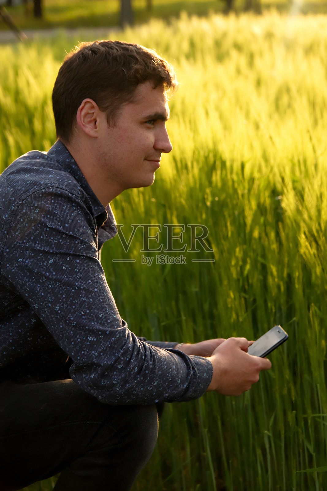夕阳下拿着手机的男人。年轻的农民在绿色的麦田里工作，用智能手机、手机检查和调整灌溉中心的枢轴洒水系统照片摄影图片