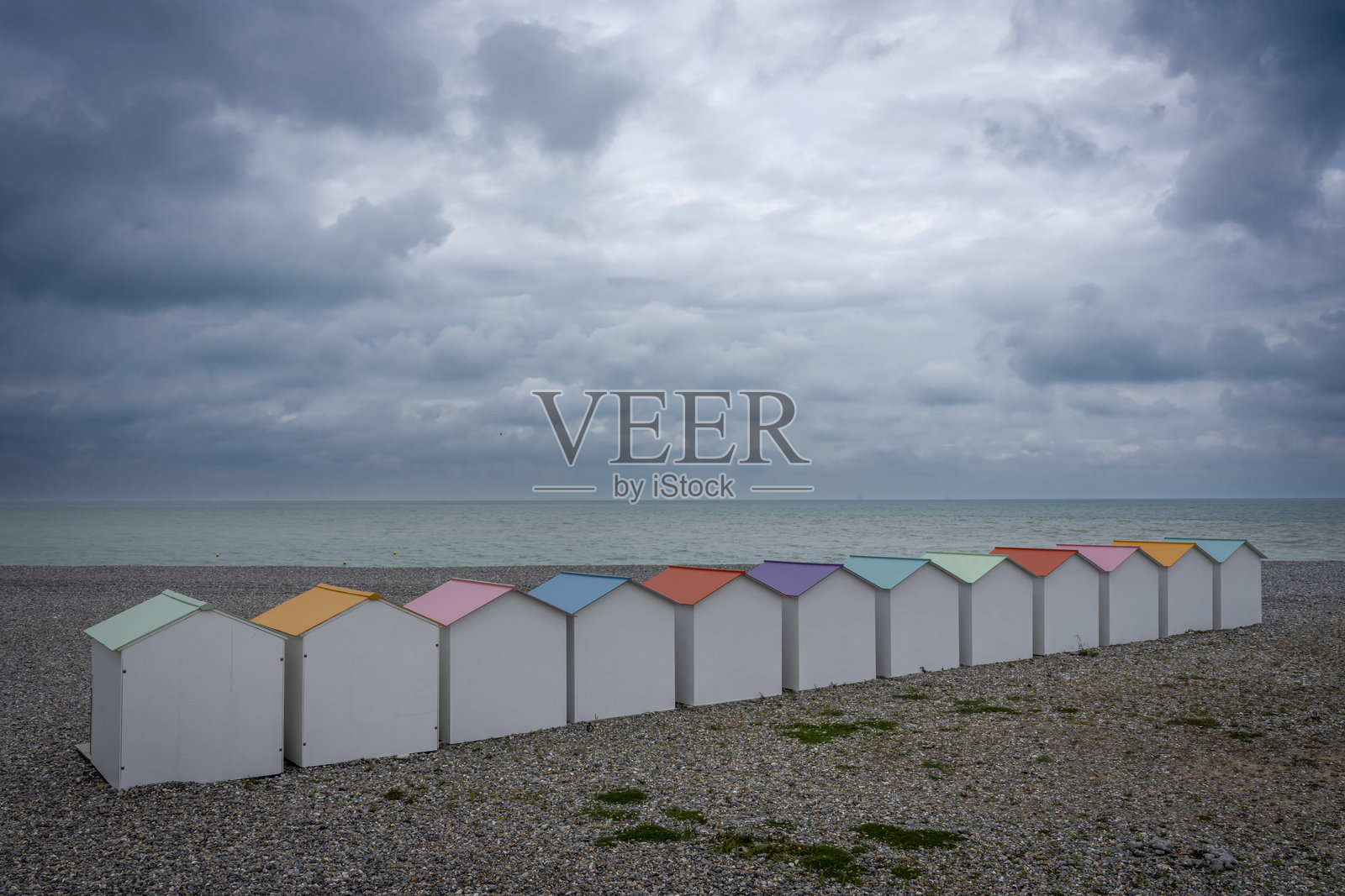 法国勒特雷波特 - 2024年9月16日：白色海滩小屋与五彩屋顶、木栈道、卵石海滩、蓝色海洋和多云的天空的景观照片摄影图片