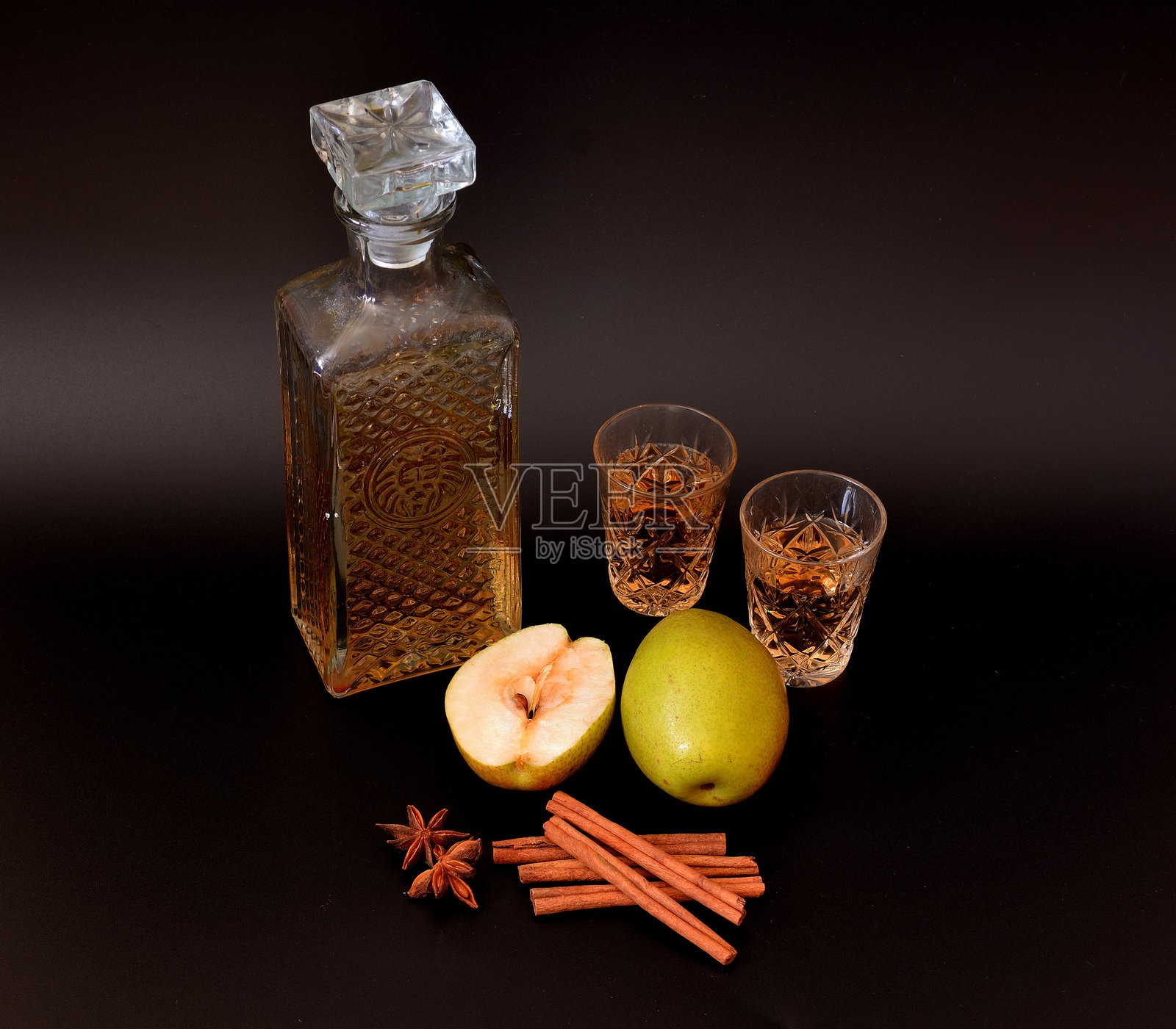 黑色背景上的梨子利口酒，加入肉桂和八角，手工制作的烈酒装在水晶酒壶中，旁边有两个酒杯，一排成熟的水果和香料。照片摄影图片