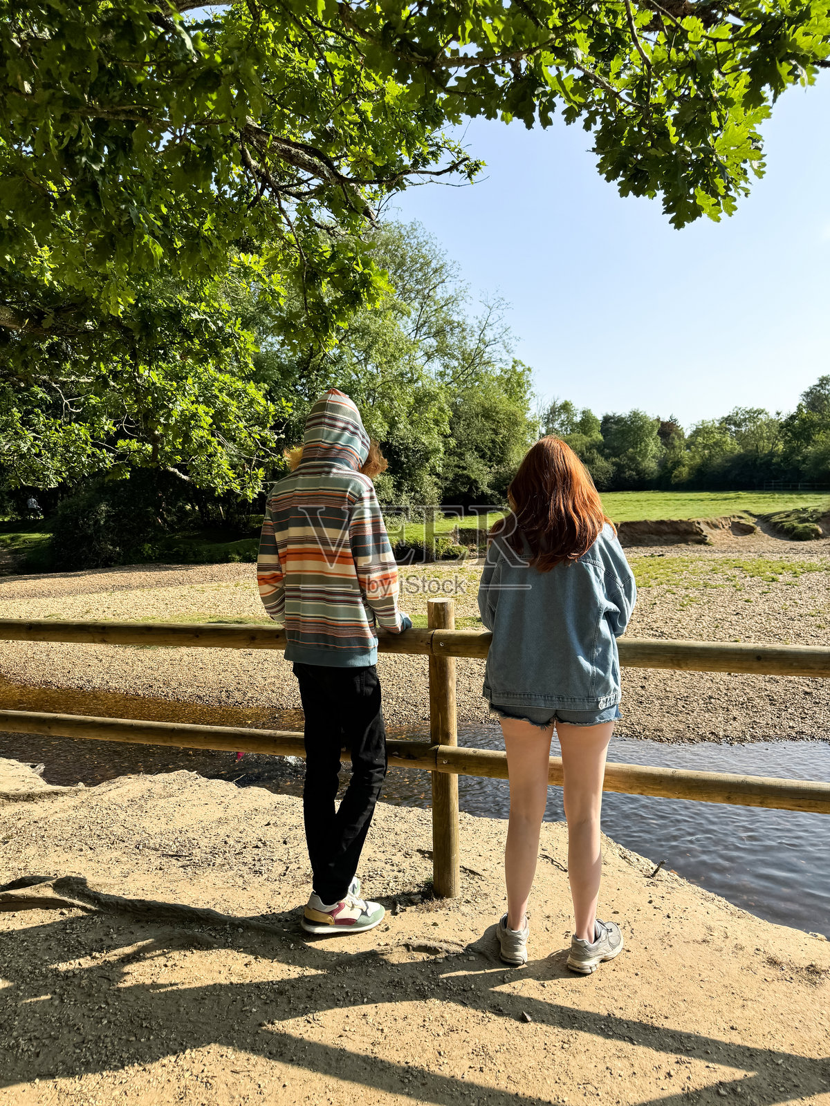 红发兄妹站在河岸的木栅栏后，河滩和流动的水，兄弟姐妹，阳光明媚的日子，背影，重点在前景。照片摄影图片