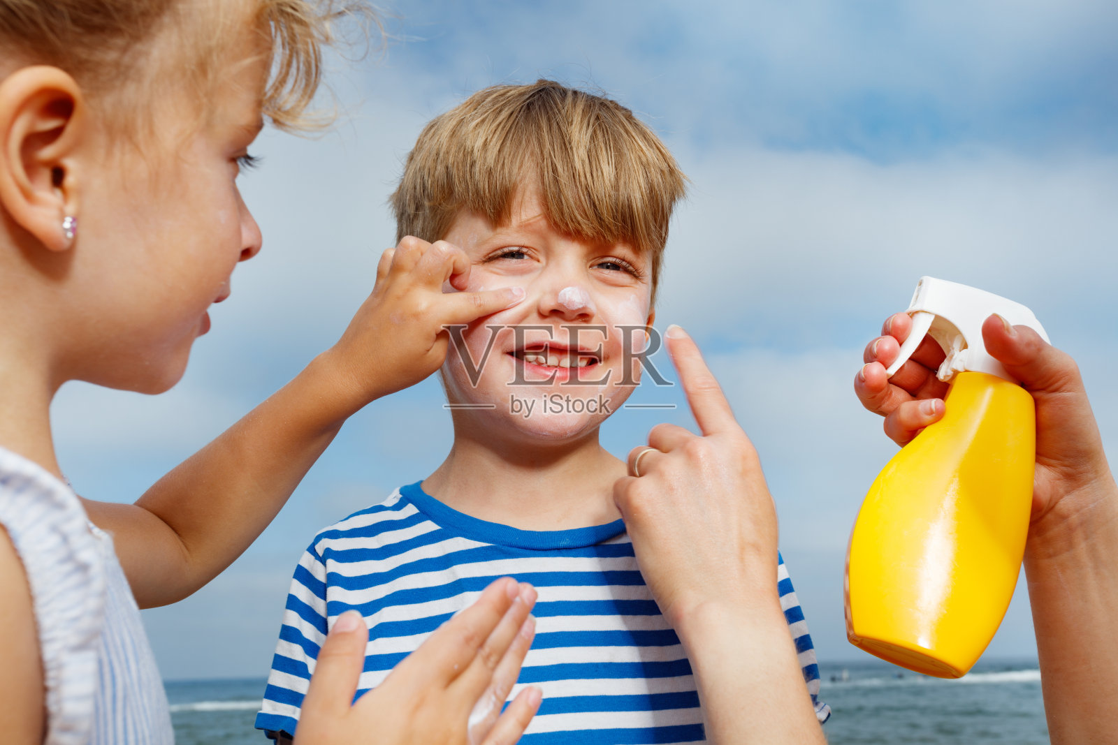 小男孩在海滩上享受姐姐为他涂抹防晒霜。照片摄影图片