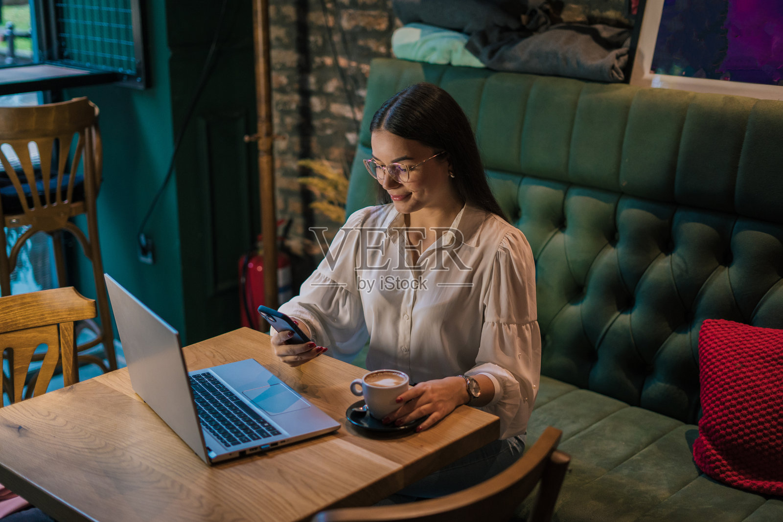 年轻女商人在咖啡馆的桌子上坐着，使用手机并在笔记本电脑上工作。照片摄影图片