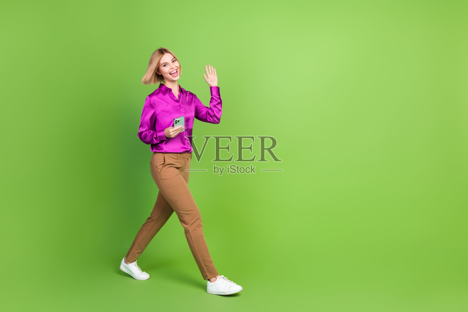 全身照，漂亮的年轻女性手持手机，走路时挥手，穿着衬衫，背景为绿色。照片摄影图片