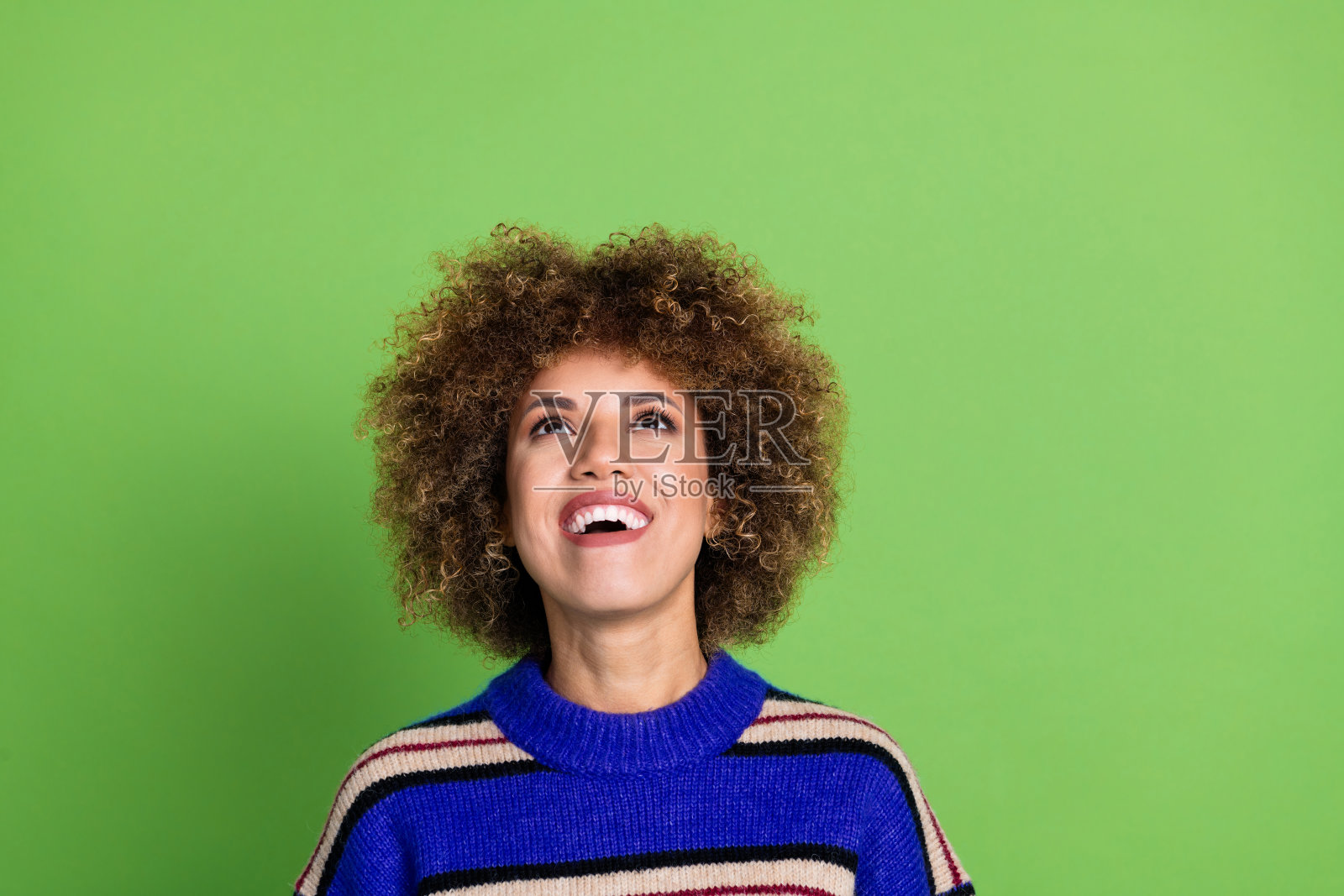 微笑露齿的女性，卷发，穿着针织毛衣，仰望空白标志，背景为绿色。照片摄影图片