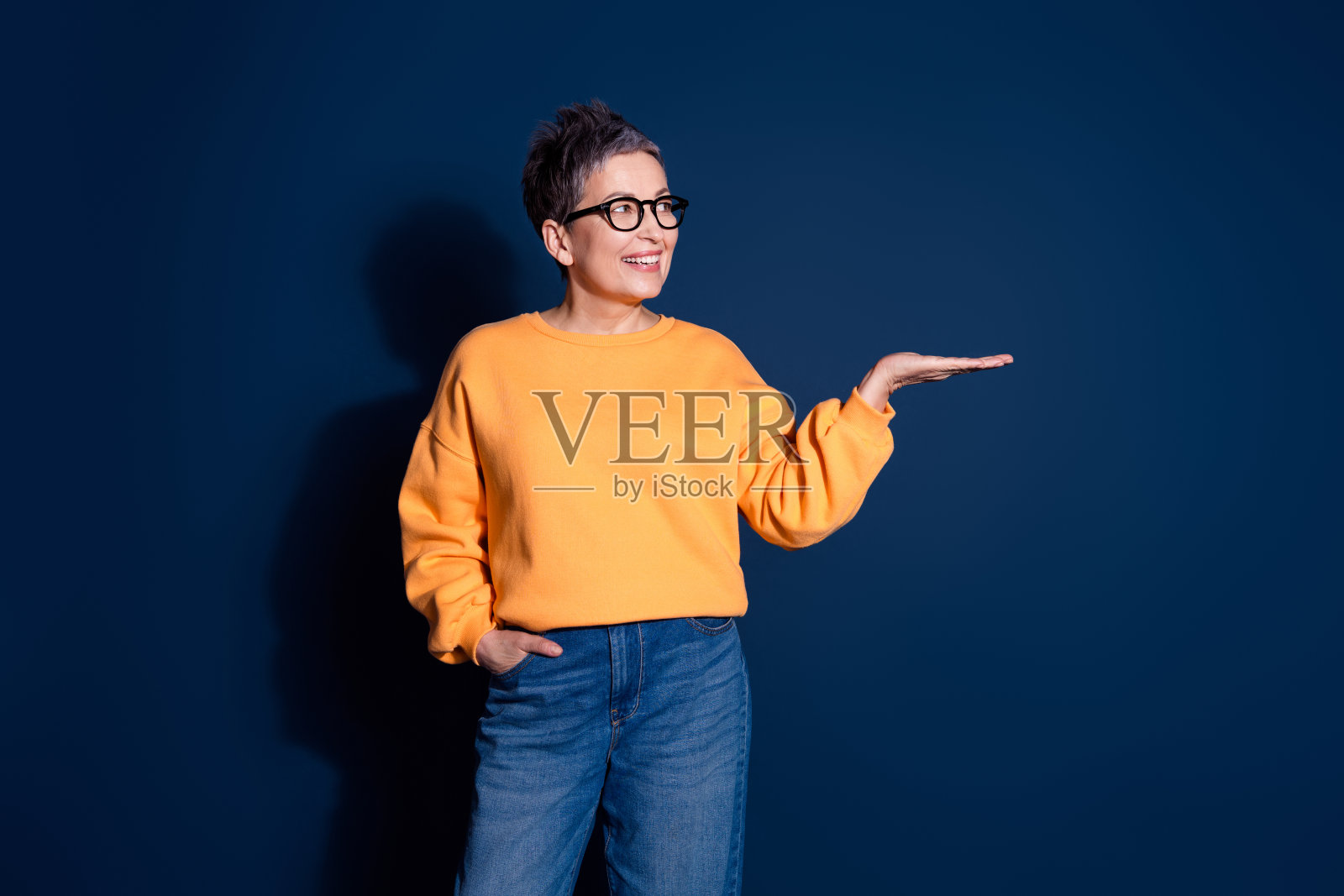 可爱的退休女士的照片肖像，穿着时尚的黄色服装，手持空白空间，背景为深蓝色。照片摄影图片