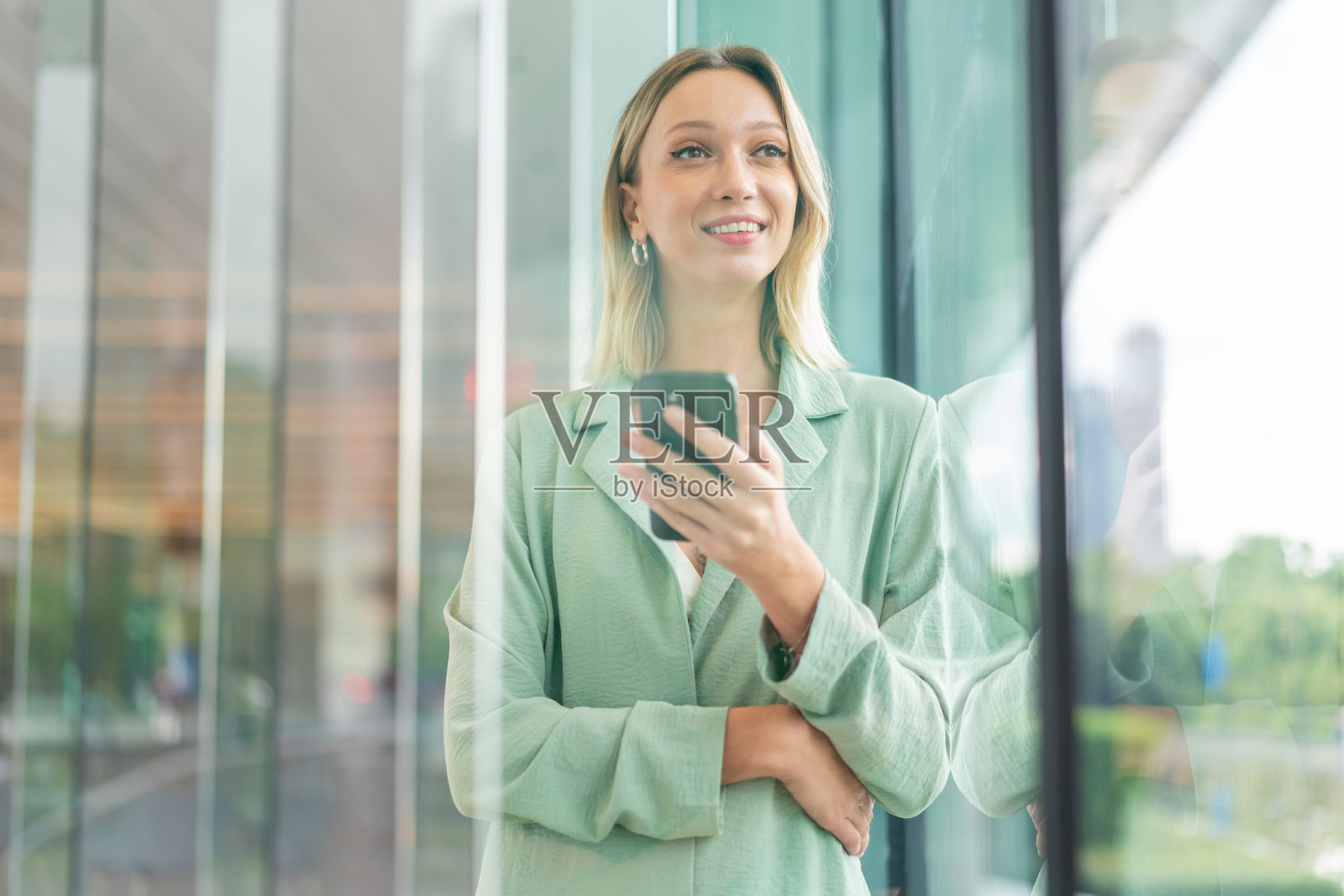 自信的金发女性穿着薄荷色西装外套，手持智能手机，靠近玻璃窗，窗外景色倒映在玻璃上。照片摄影图片