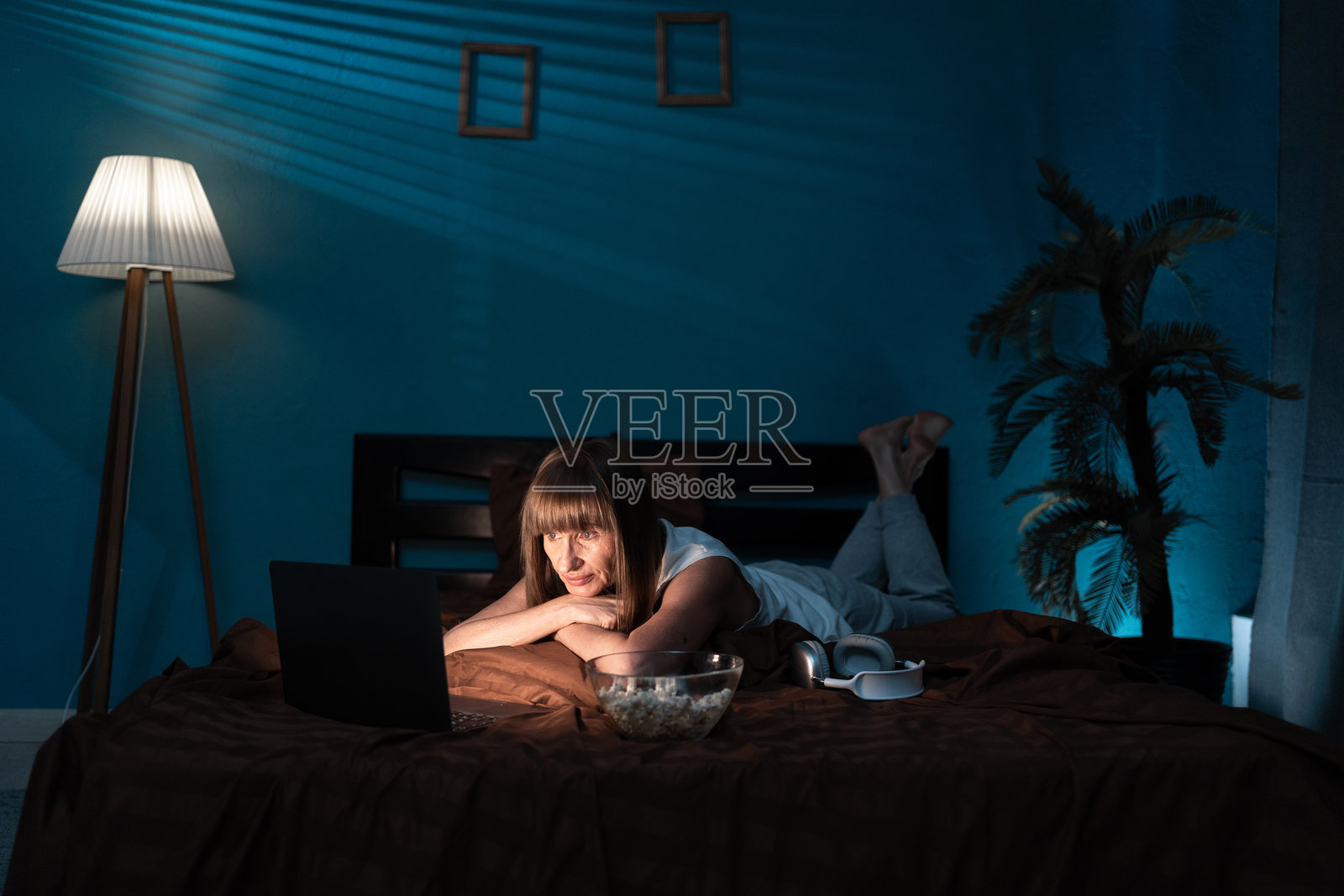 中年女性在夜晚躺在床上观看一部有趣的电影，边吃爆米花。照片摄影图片