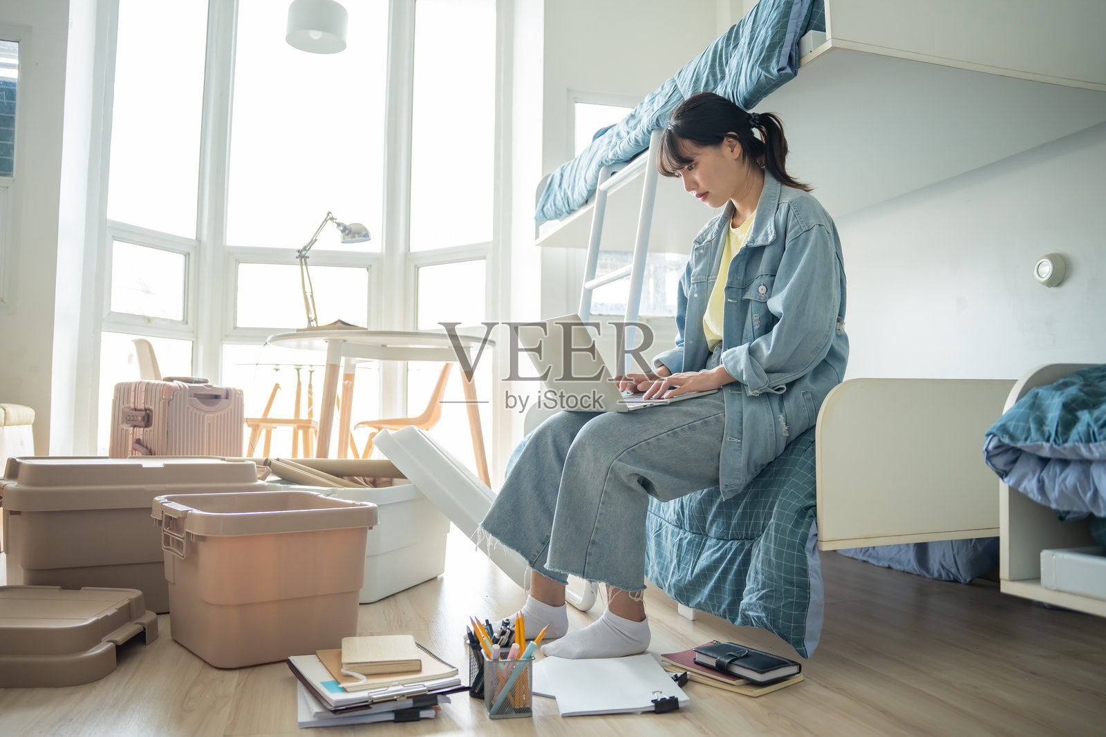 亚洲女孩坐在床上，正在笔记本电脑上工作，房间很乱。照片摄影图片