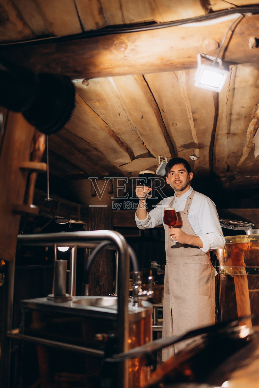 英俊的有胡子的酿酒专家手持啤酒杯，凝视着杯中的啤酒。酿酒工人站在酿酒设备旁，检查啤酒杯。生产控制的概念。照片摄影图片