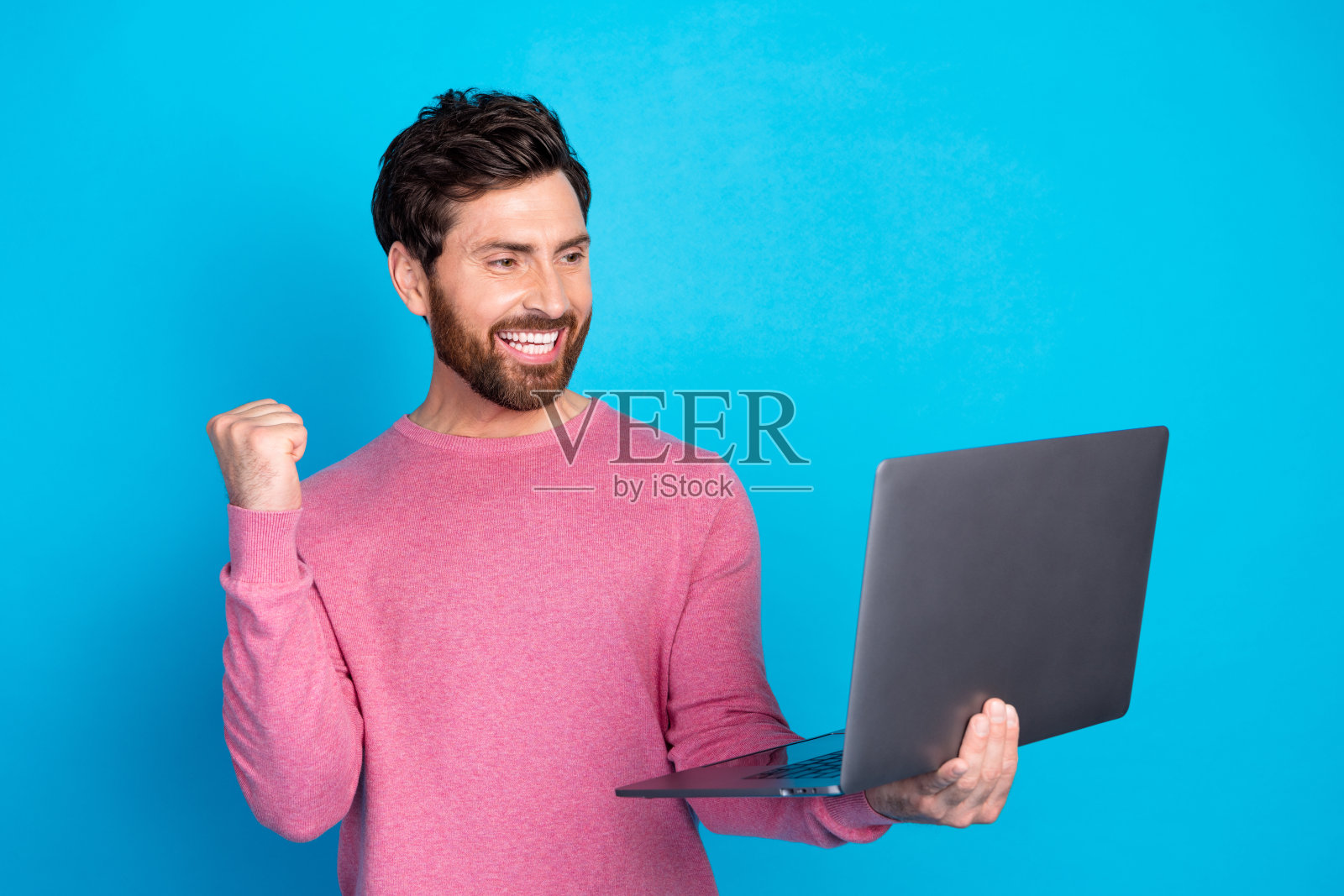 自信的络腮胡男子在使用笔记本电脑时，挥拳庆祝成功，背景为蓝色照片摄影图片
