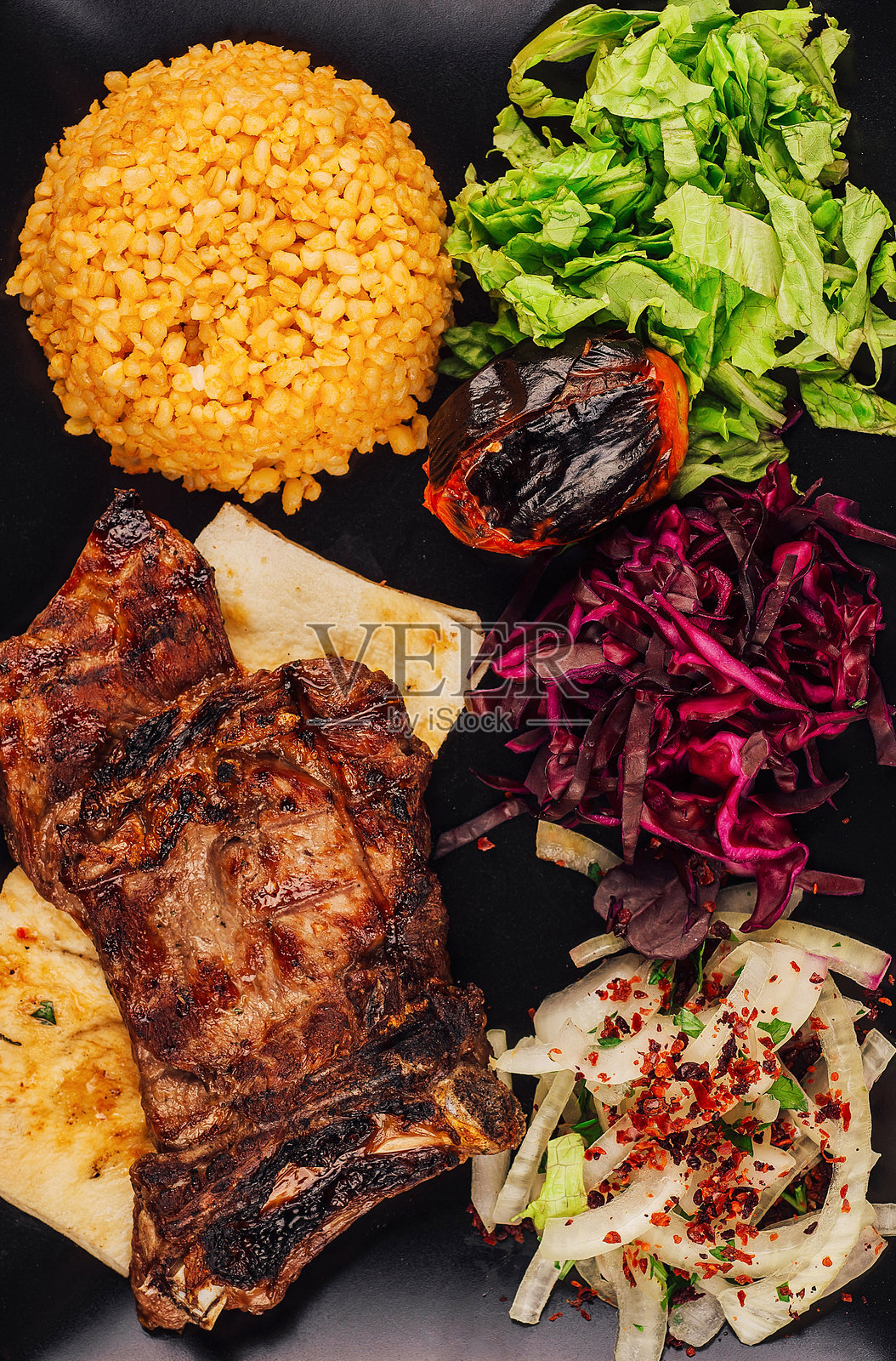 黑色盘子上摆放着烤羊肋骨，配有粗麦粉、沙拉和烤番茄。照片摄影图片