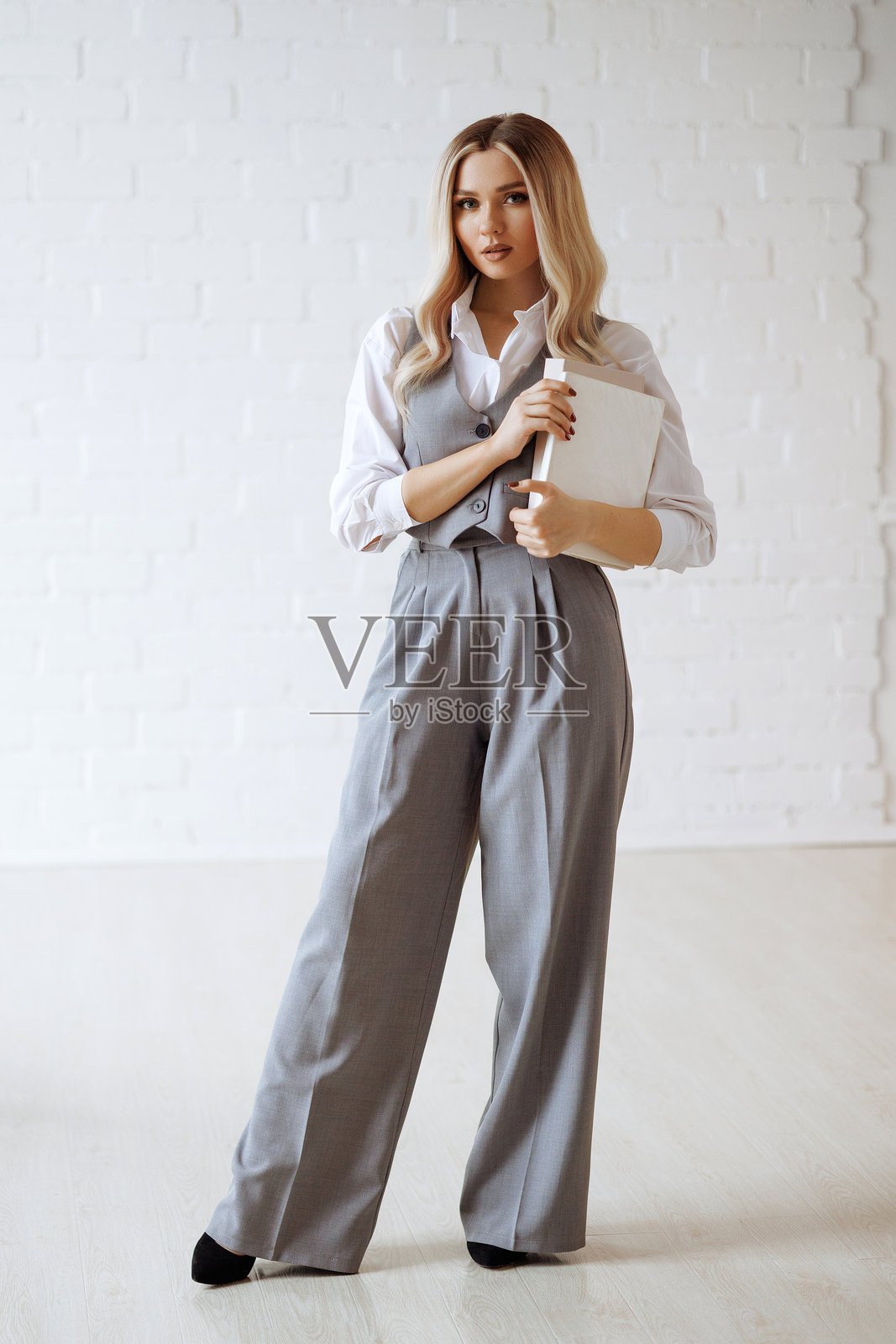 穿着灰色西装和白色衬衫的金发女子拿着一本书站在白色背景上照片摄影图片