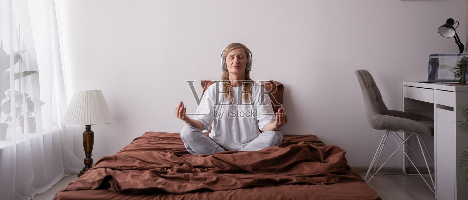 早晨冥想概念。老年女性坐在床上以莲花坐姿冥想，晨光洒在她的卧室里。照片摄影图片