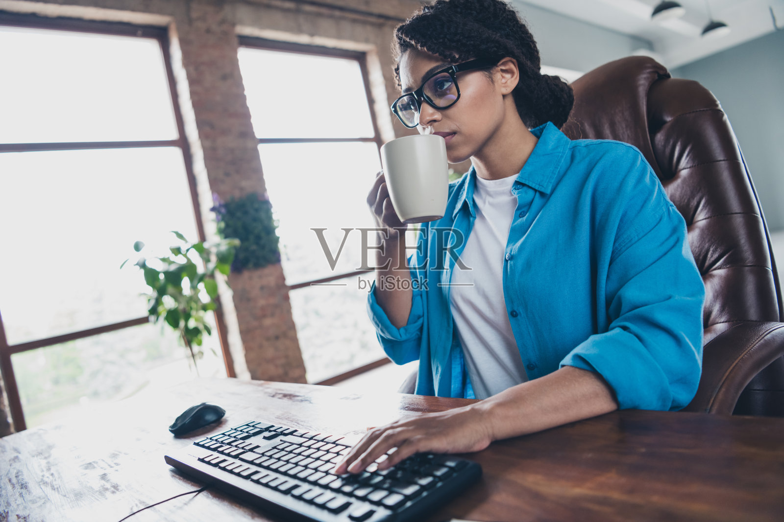 漂亮年轻女孩在电脑前打字，喝咖啡，穿着蓝色衬衫，坐在共享办公空间，程序员，成功，明亮的办公室。照片摄影图片