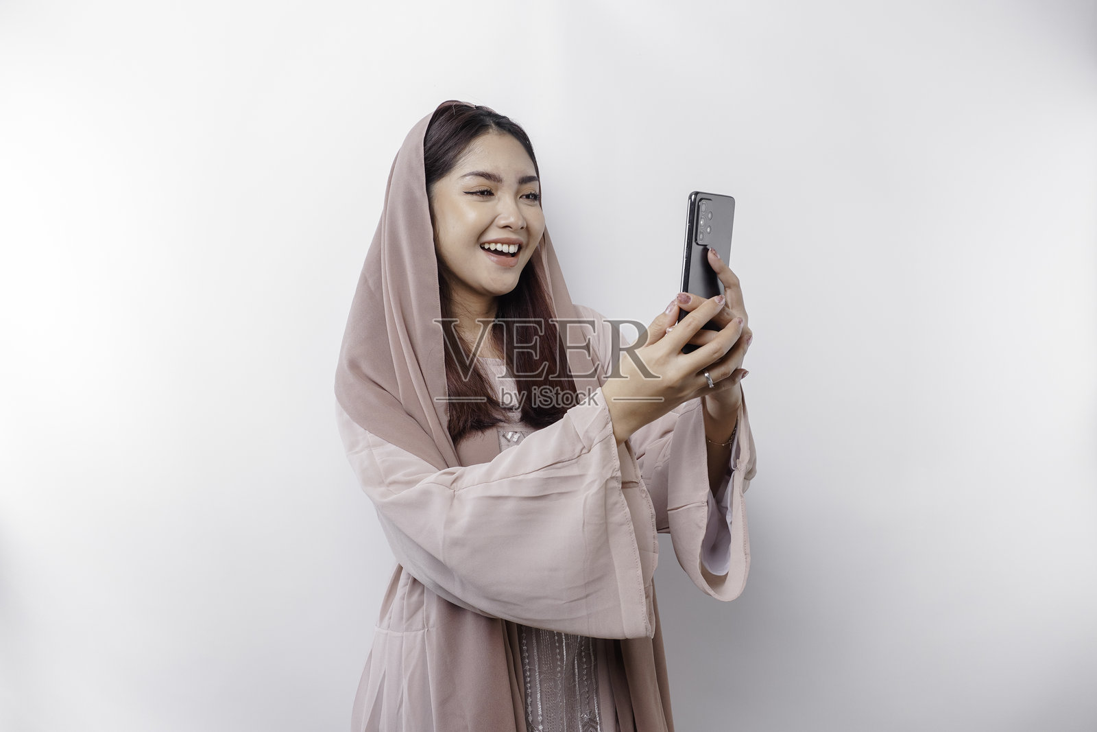 一位快乐的亚洲穆斯林女性，戴着头巾，手里拿着手机。照片摄影图片