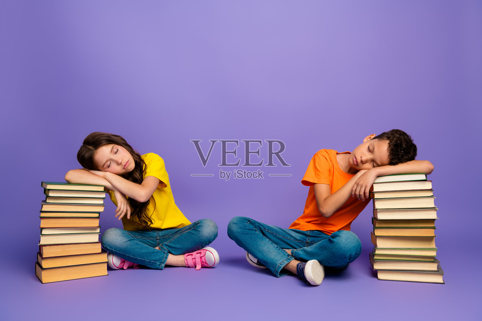 两位可爱的小孩穿着时尚的橙黄色衣服，疲惫地躺在书本上，背景为紫色。照片摄影图片