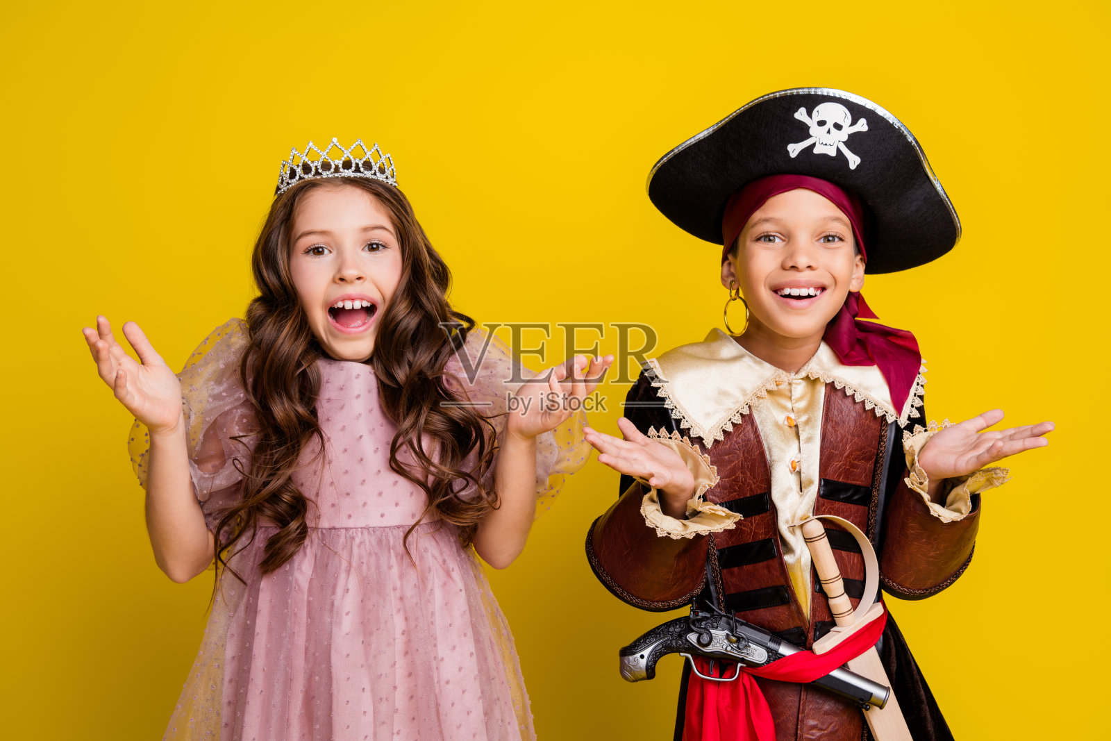 两个小孩举起手，张嘴欢呼，穿着万圣节海盗公主服装，背景为黄色。照片摄影图片