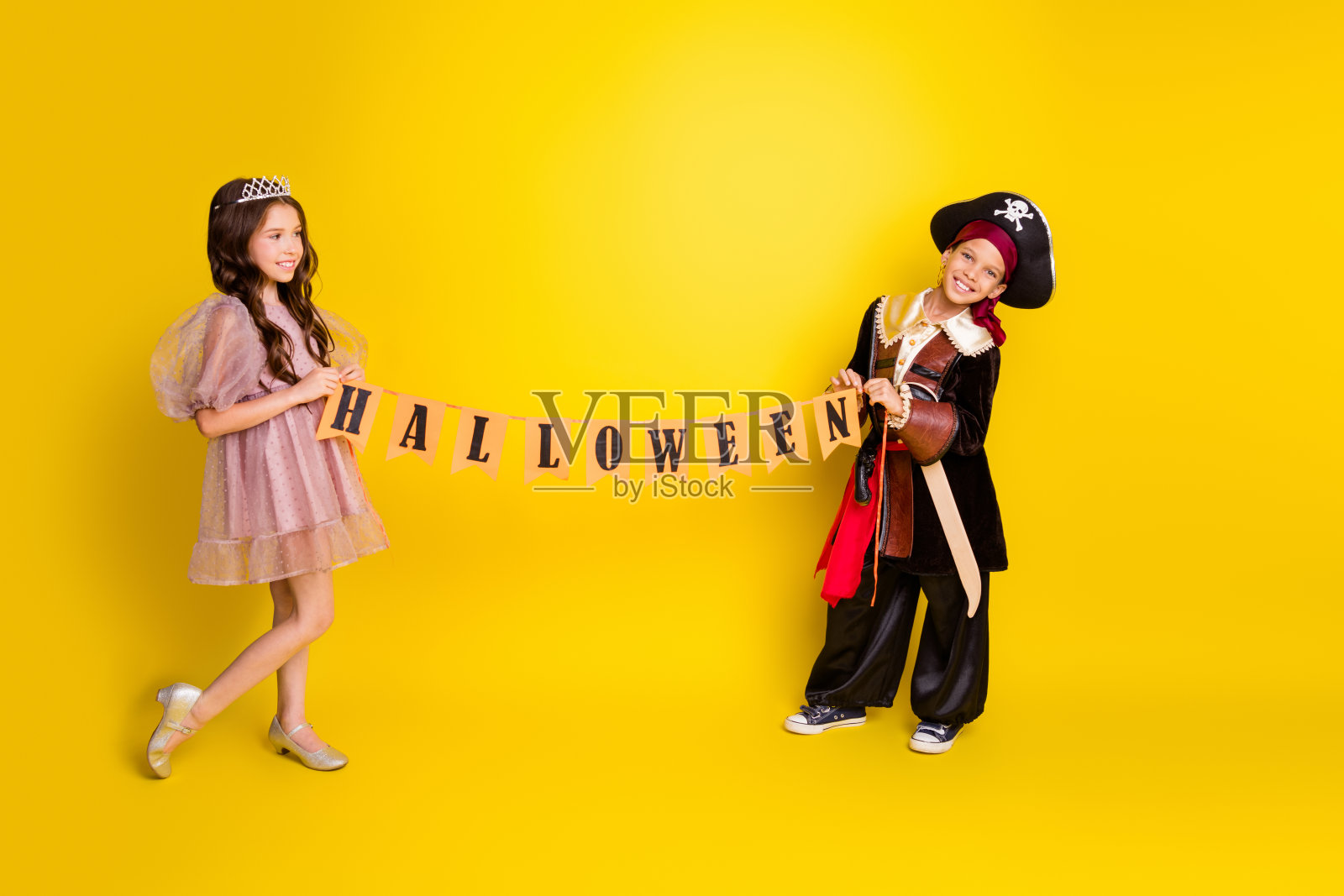 两个孩子穿着公主和海盗服装，手持万圣节旗帜，完整身影肖像，背景为黄色。照片摄影图片