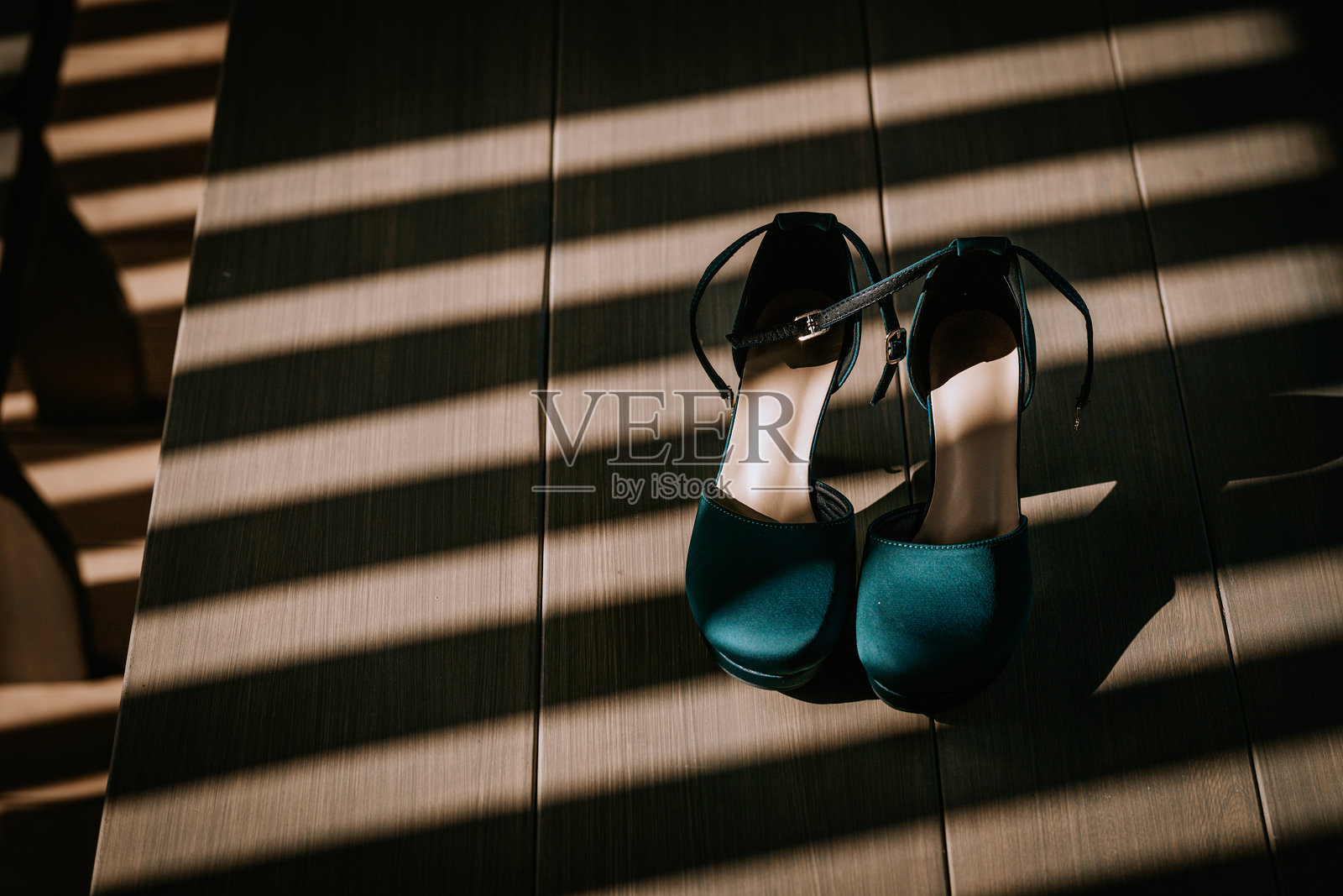 一双优雅的青色高跟鞋放在木地板上，阳光透过条纹状的阴影照射，营造出戏剧性和时尚感。照片摄影图片