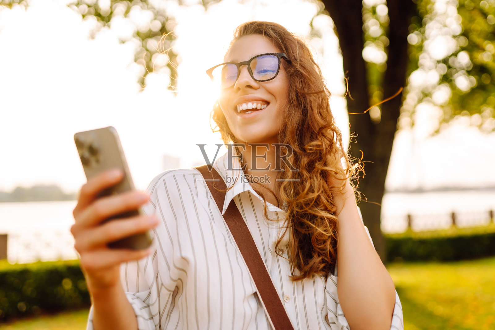 一位美丽的卷发女性在阳光明媚的公园里微笑着使用智能手机，身旁是水面，正值黄金时刻。照片摄影图片