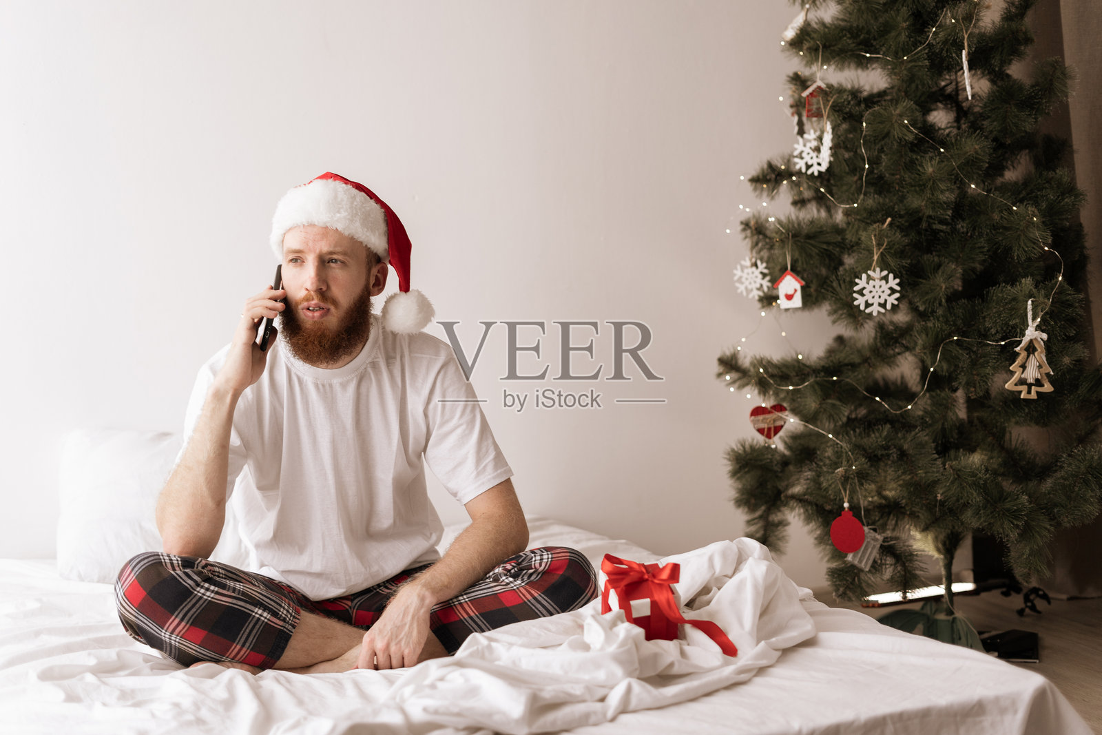 身穿舒适睡衣的年轻男人在卧室床上用手机，背景是圣诞树照片摄影图片