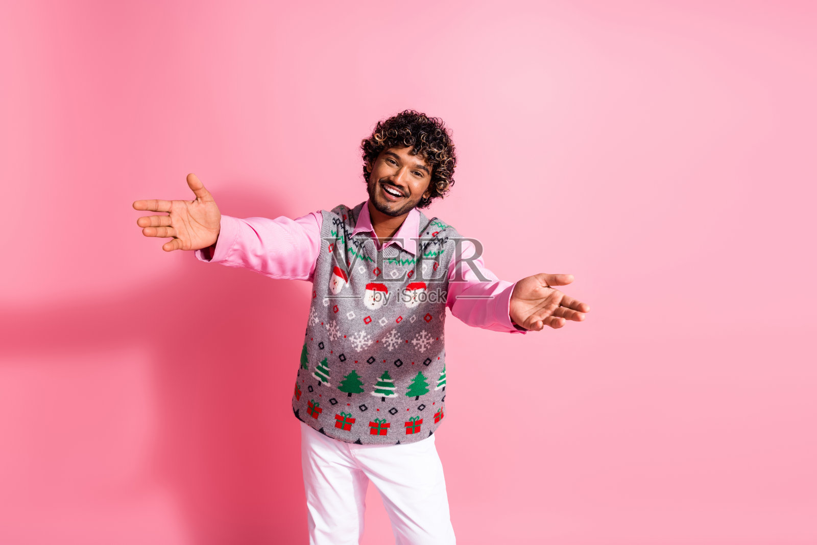 年轻男子张开双臂欢迎，身穿圣诞背心，粉色背景照片摄影图片