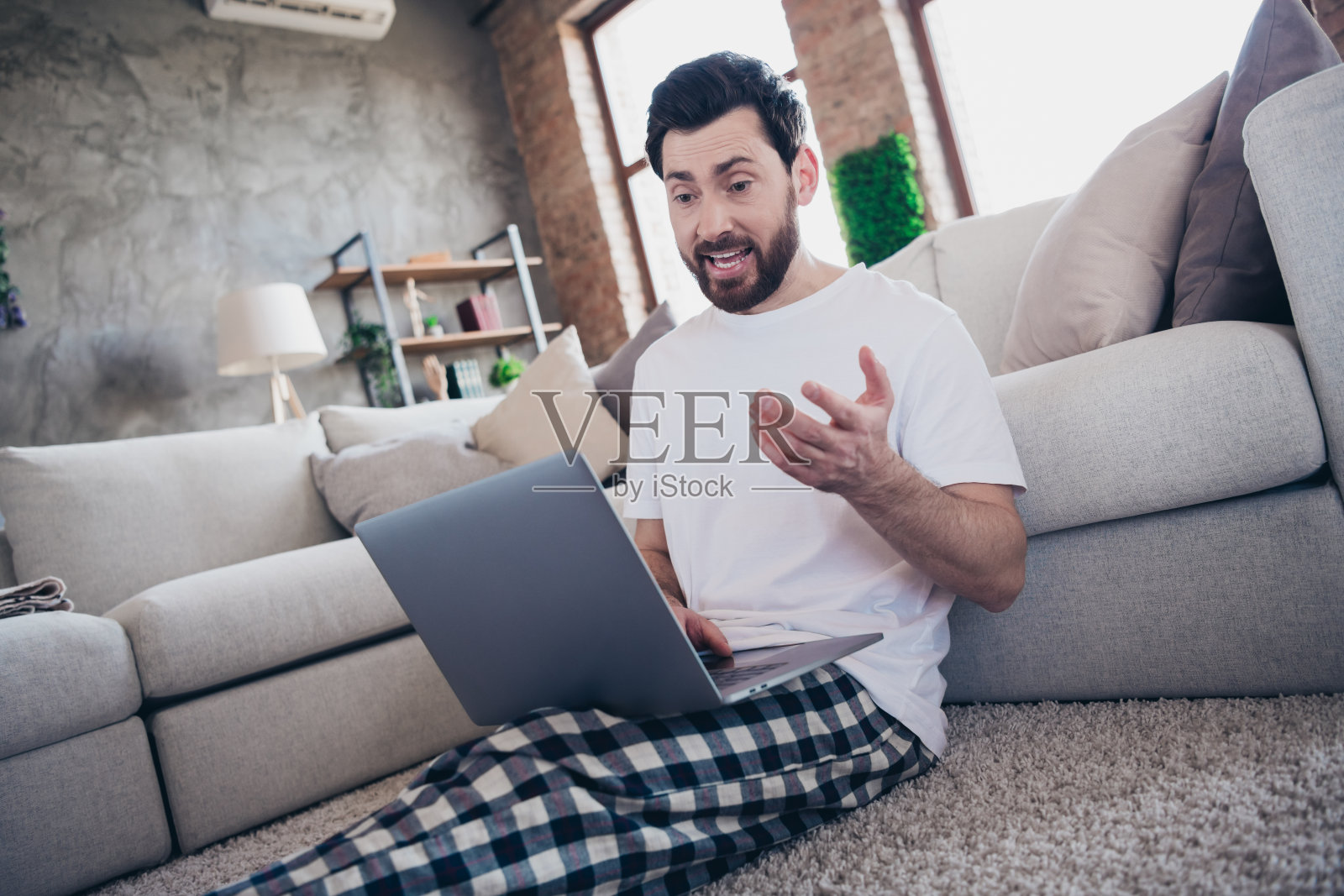 穿着睡衣的迷人胡子男在家工作，使用现代设备，坐在室内的客厅里。照片摄影图片