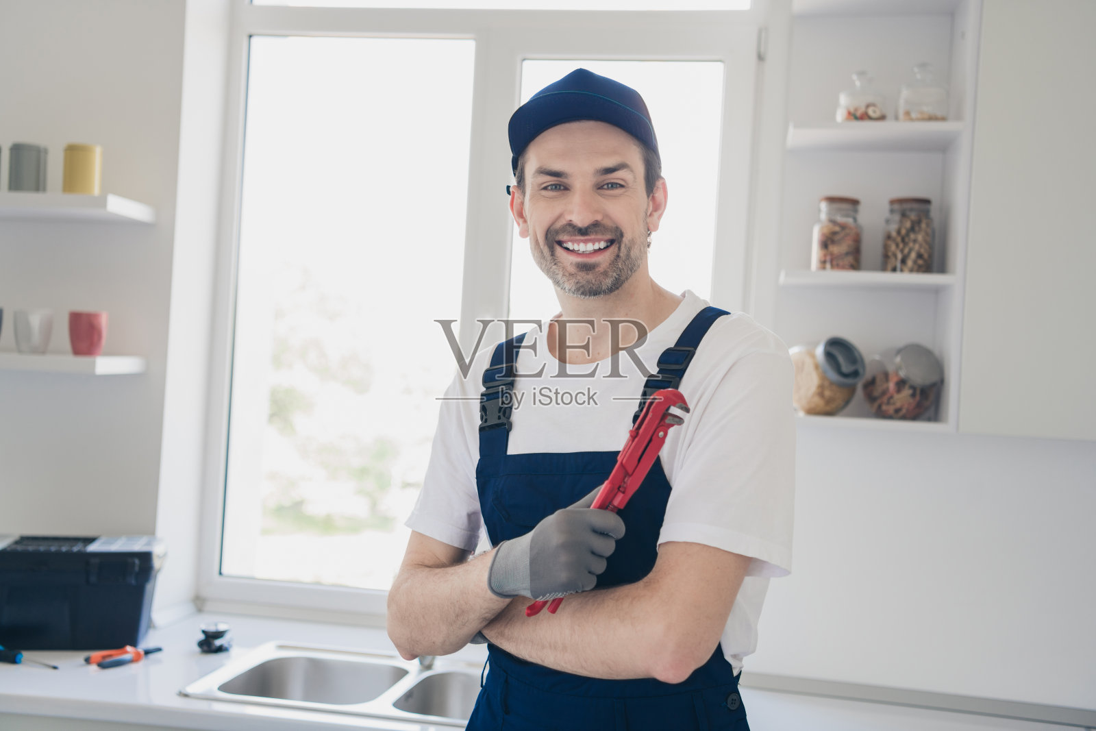 微笑的修理工穿着工装，手持扳手，站在明亮现代的厨房中，准备进行家庭装修或管道维修。照片摄影图片