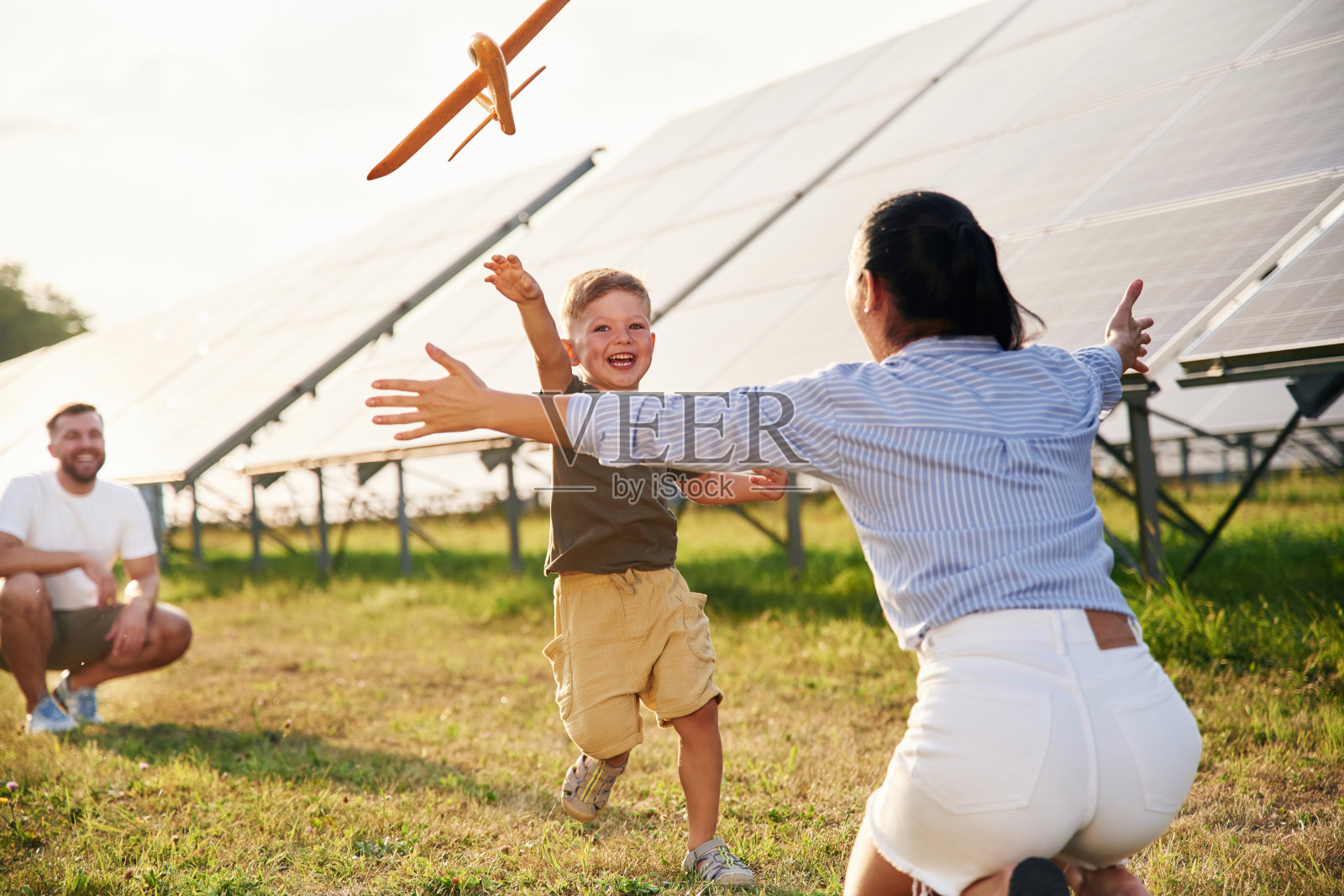 奔向拿着玩具飞机的父母。父亲、母亲和小儿子在太阳能电池板附近照片摄影图片