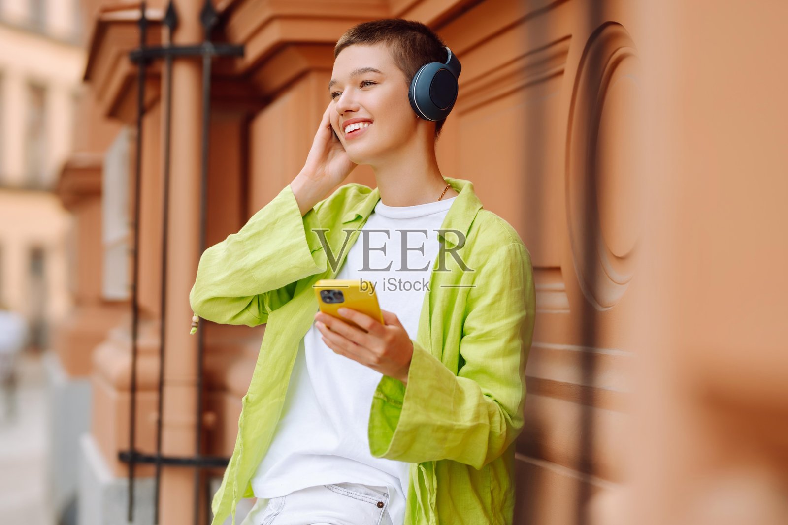 年轻女性穿着明亮的绿色夹克，在城市环境中戴着耳机享受音乐，同时使用智能手机。照片摄影图片