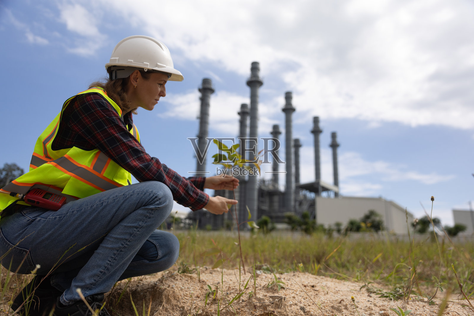 一位聪明的女性项目负责人正在手动种植可再生资源树木，以减少污染，手中展示环境、社会和可持续商业治理的ESG图标，旁边是一座火力发电厂。照片摄影图片
