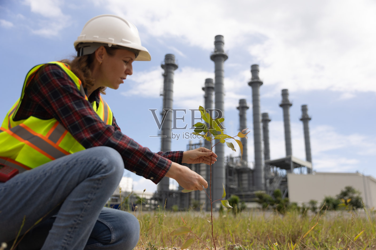 一位聪明的女性项目负责人正在手动种植树木，以减少污染，手中展示可再生资源的ESG图标概念，旁边是一座火力发电厂。照片摄影图片