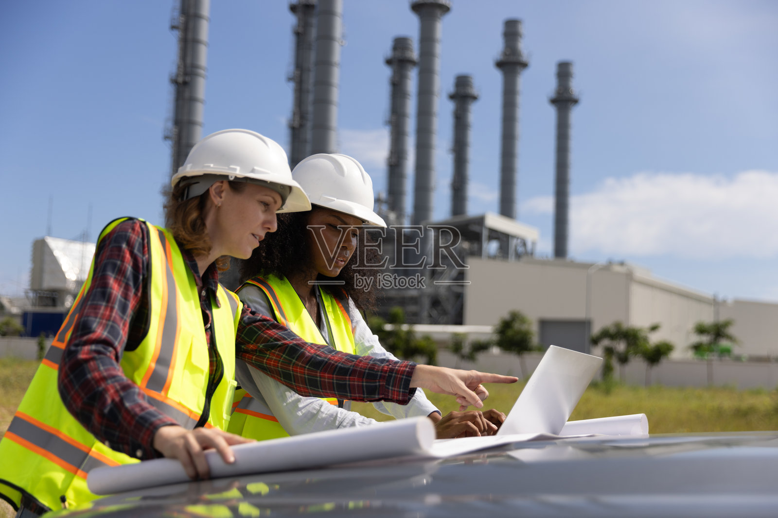 在化工厂，两位能干的女建筑工程师正在用蓝图研究汽车引擎盖和后备箱。照片摄影图片