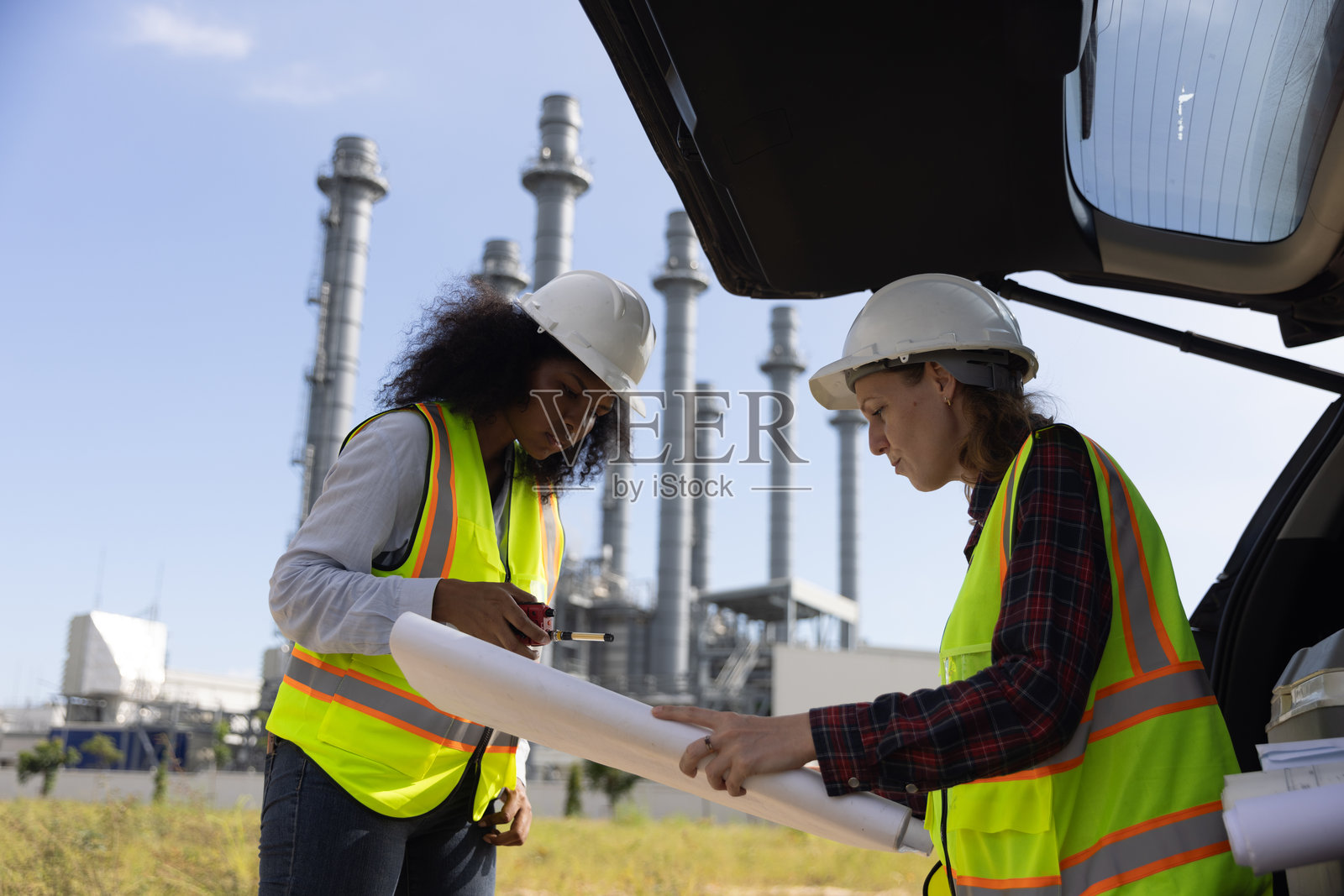 在化工厂，两位能干的女建筑工程师正在用蓝图研究汽车引擎盖和后备箱。照片摄影图片