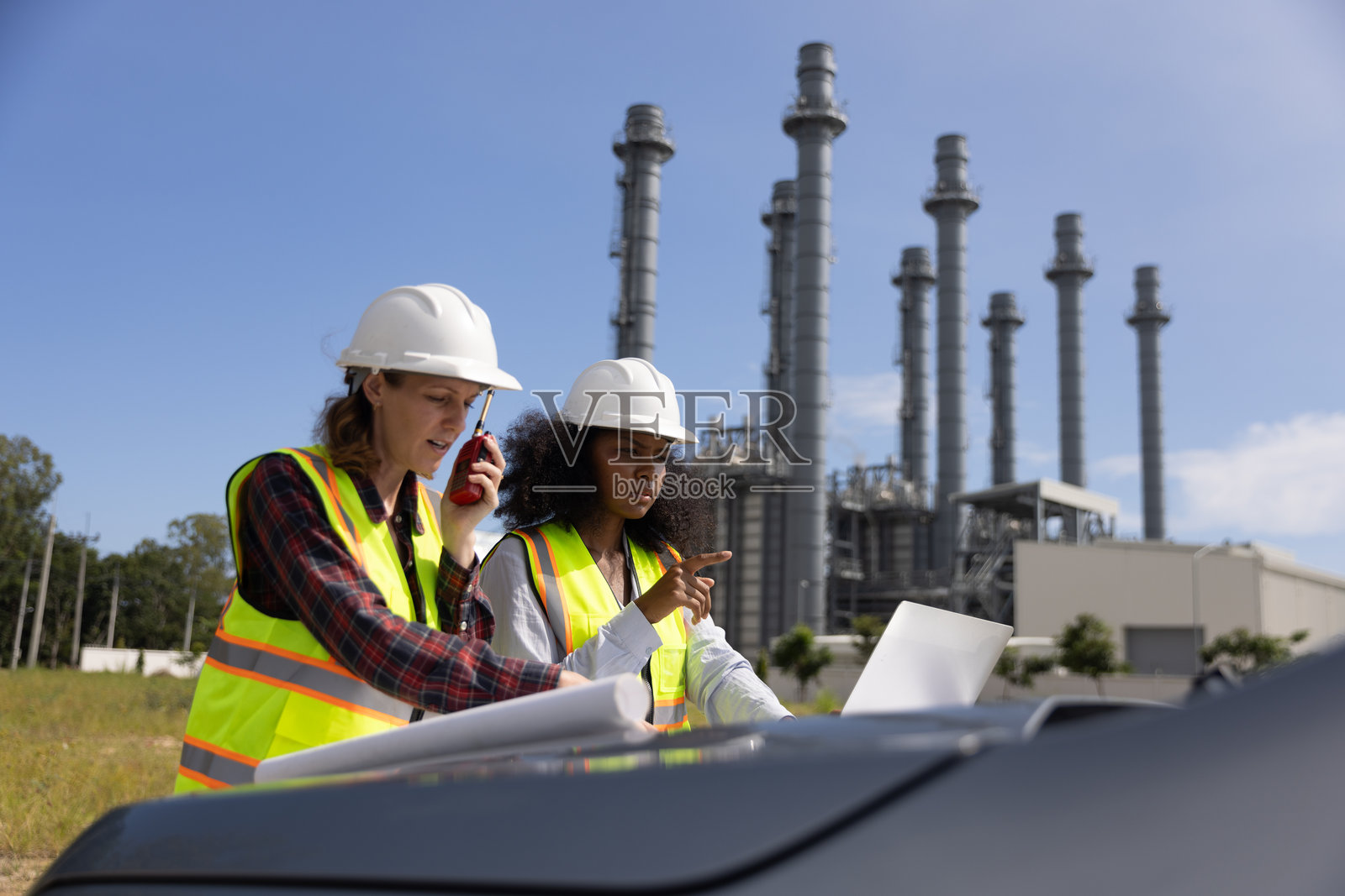 在化工厂，两位能干的女建筑工程师正在用蓝图研究汽车引擎盖和后备箱。照片摄影图片