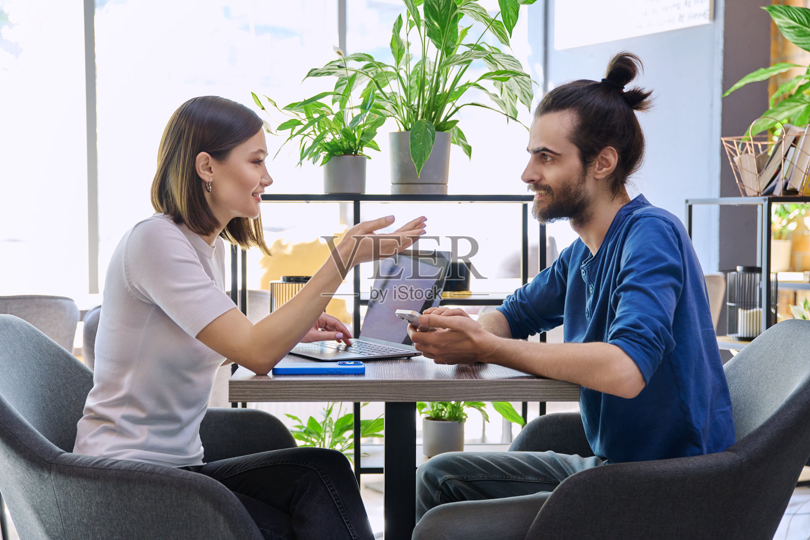年轻男女坐在共享办公空间的桌子旁，使用笔记本电脑和智能手机，正在交谈。照片摄影图片