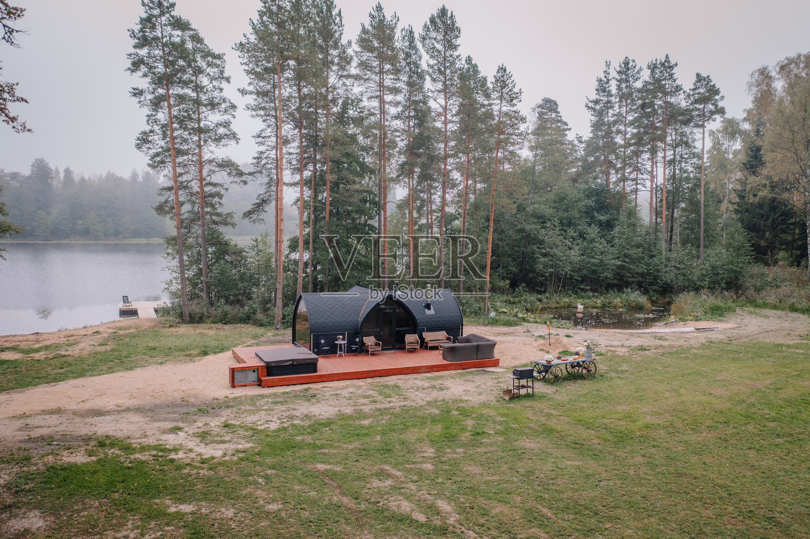 一座现代化的小屋，拥有弯曲的屋顶、木质甲板、热水浴缸和户外座位，坐落在湖边，四周环绕着树木，环境宁静自然。照片摄影图片
